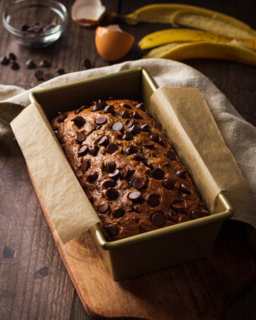 Freshly baked chocolate chip banana bread in a loaf pan with scattered chocolate chips on top sits on wooden board.