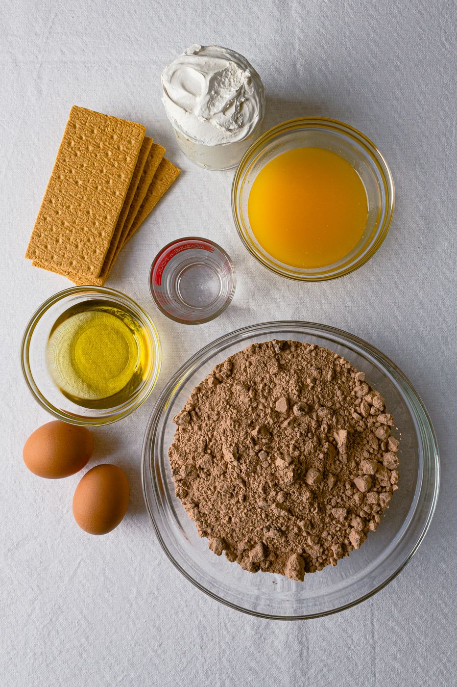Ingredients for dessert including graham crackers, whipped cream, melted butter, oil, eggs, and chocolate powder in bowls.
