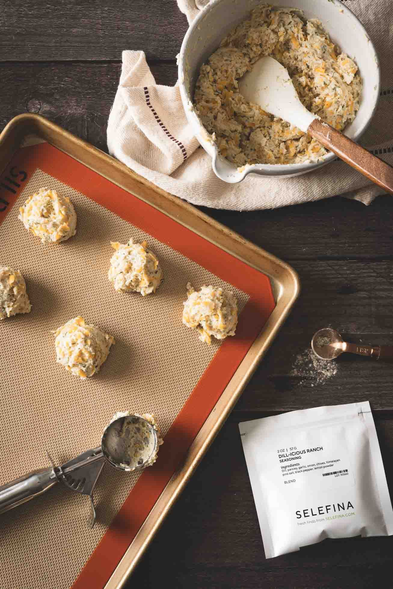Biscuit dough balls arranged on a baking sheet with mixing bowl, scoop, and Seleflina package on dark wooden surface.