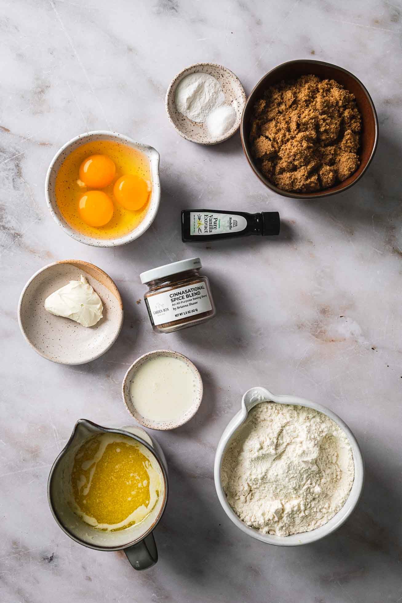 Baking ingredients including brown sugar, eggs, flour, and melted butter arranged on marble countertop with measuring bowls.