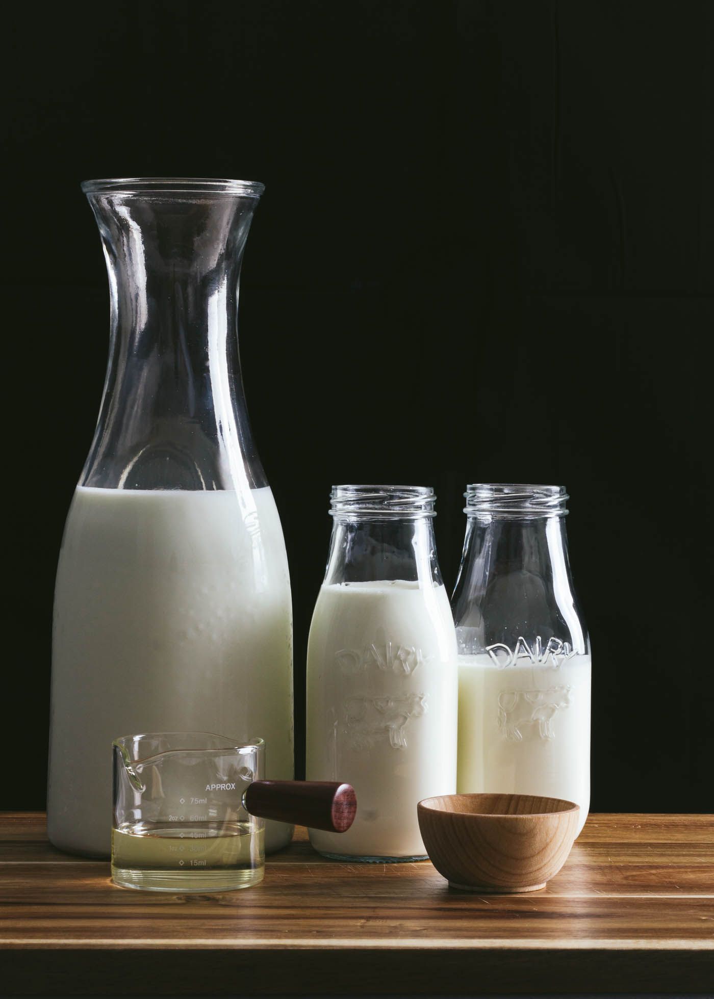 Glass bottles of fresh white milk arranged on rustic wooden table against dark background with small measuring bowl.
