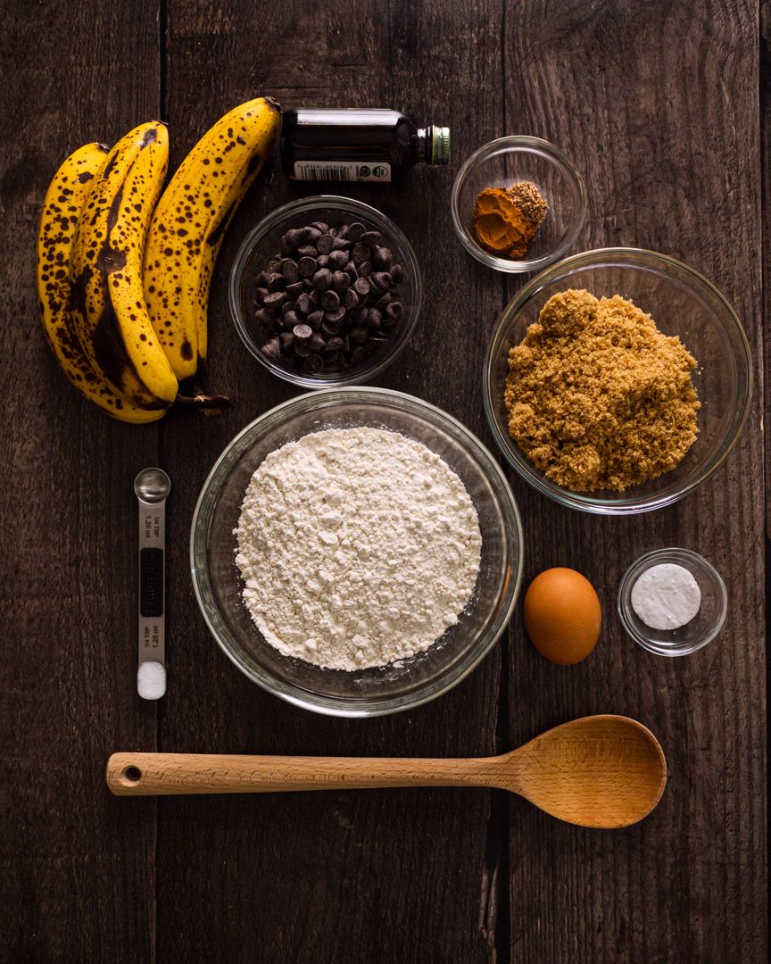Baking ingredients including ripe bananas, flour, brown sugar, chocolate chips, and an egg arranged on dark wooden surface.