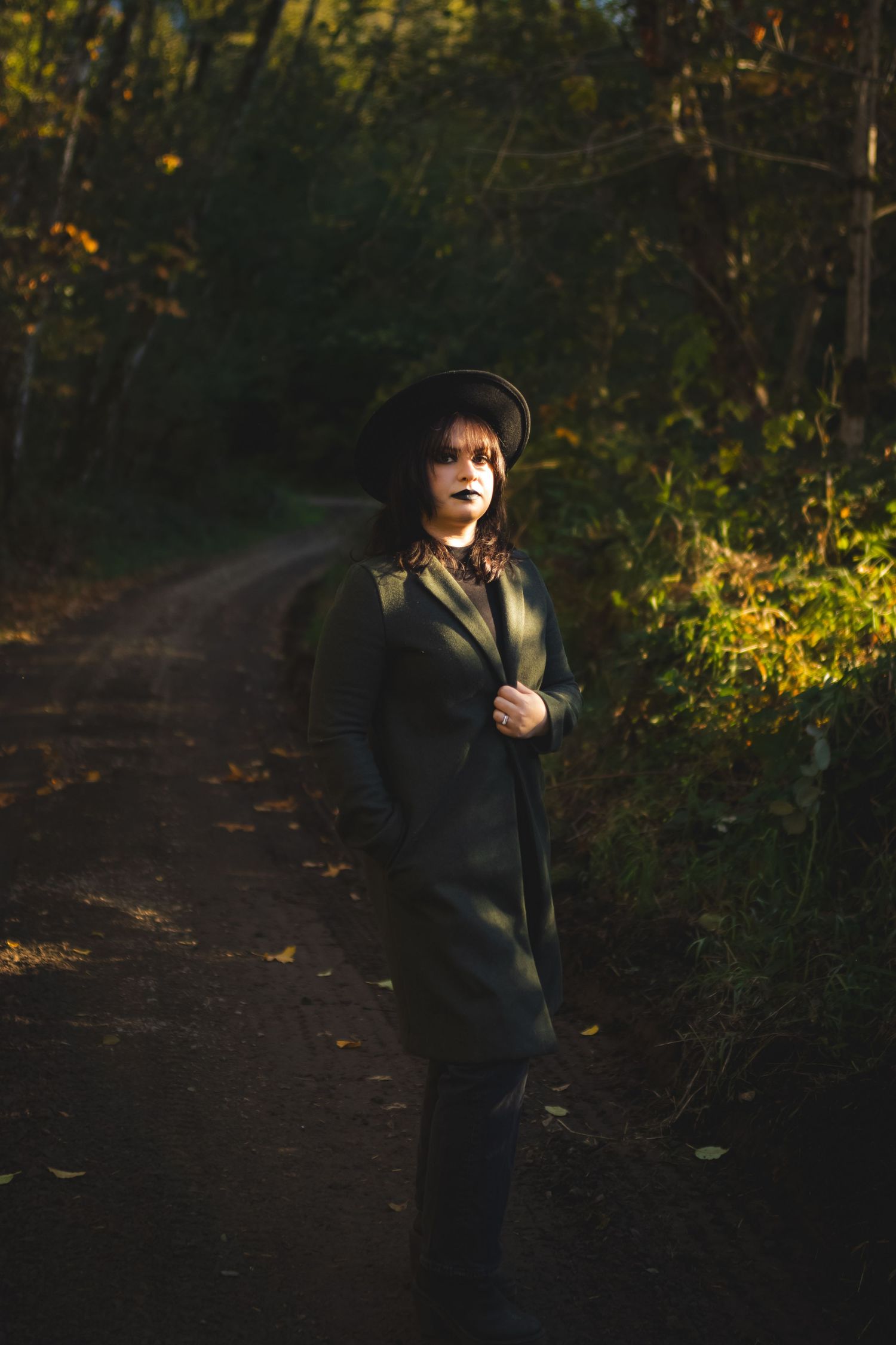 Woman wearing a dark coat and hat standing on a wooded path during a moody portrait session in Big Bear California with soft natural light