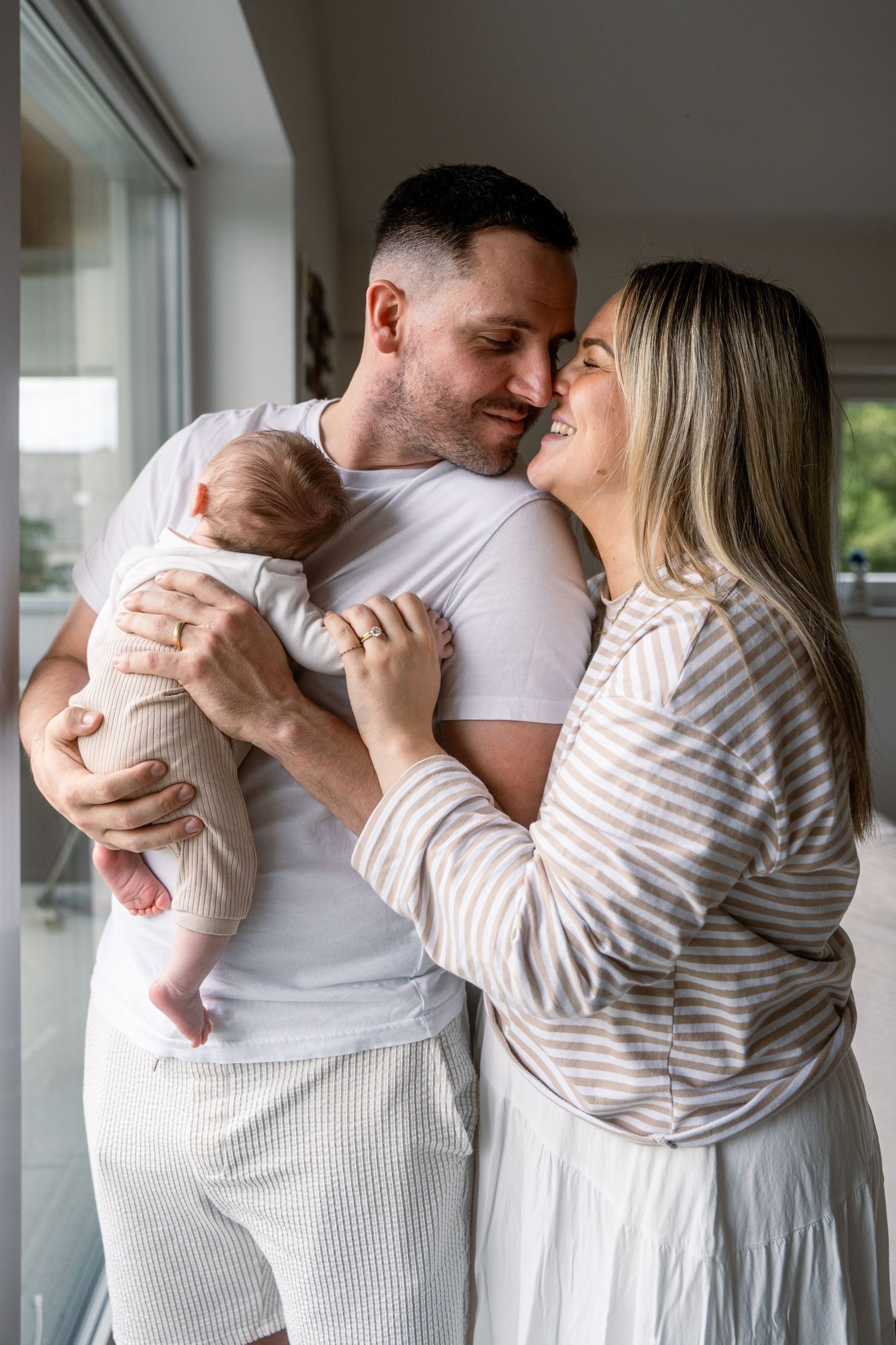 A smiling couple touches noses in a bright room while the father cradles their sleeping newborn baby in his arms.