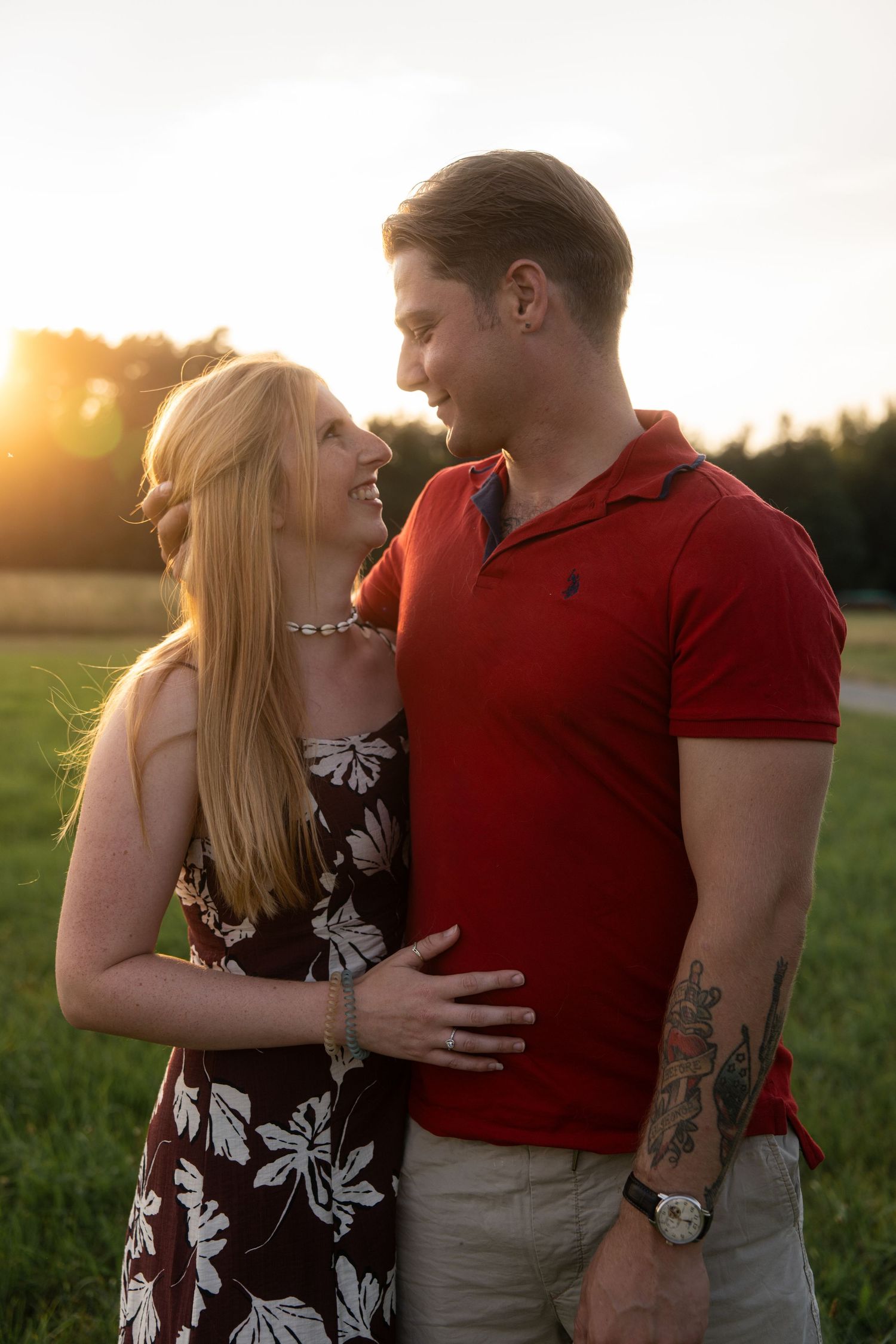 Smiling couple embracing in a grassy field at sunset, the woman in a floral dress and the man in a red polo with arm tattoos.