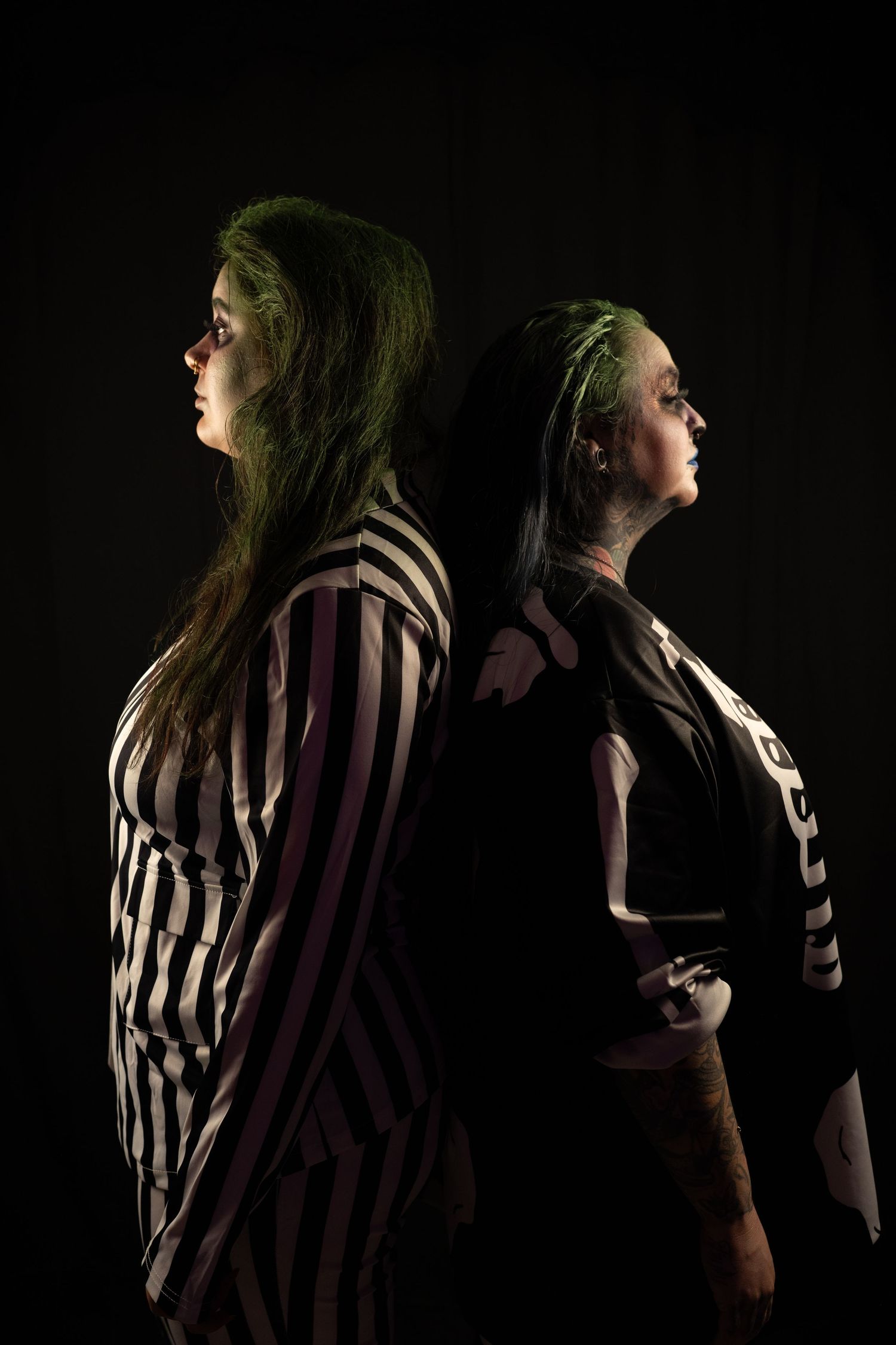 Two women stand back to back in profile, one in a striped suit, in a dramatic low-key studio portrait against a black background.