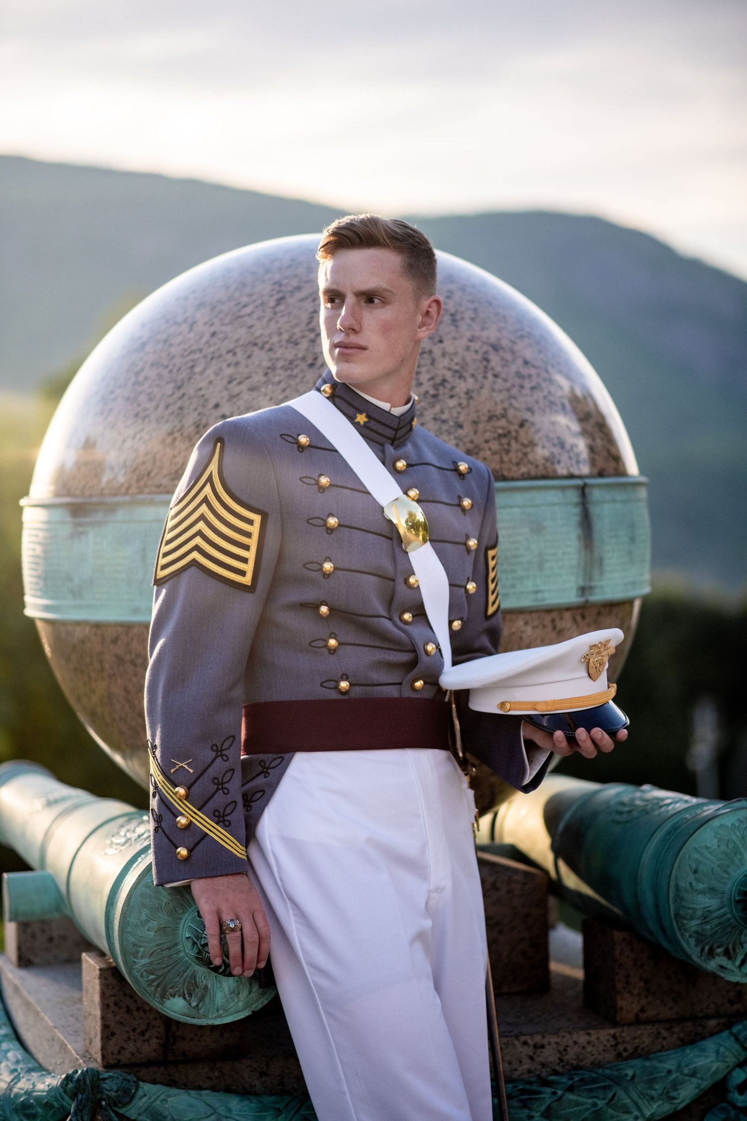 A young cadet in a gray dress uniform with gold buttons holds a white cap beside a large stone sphere and green cannons.