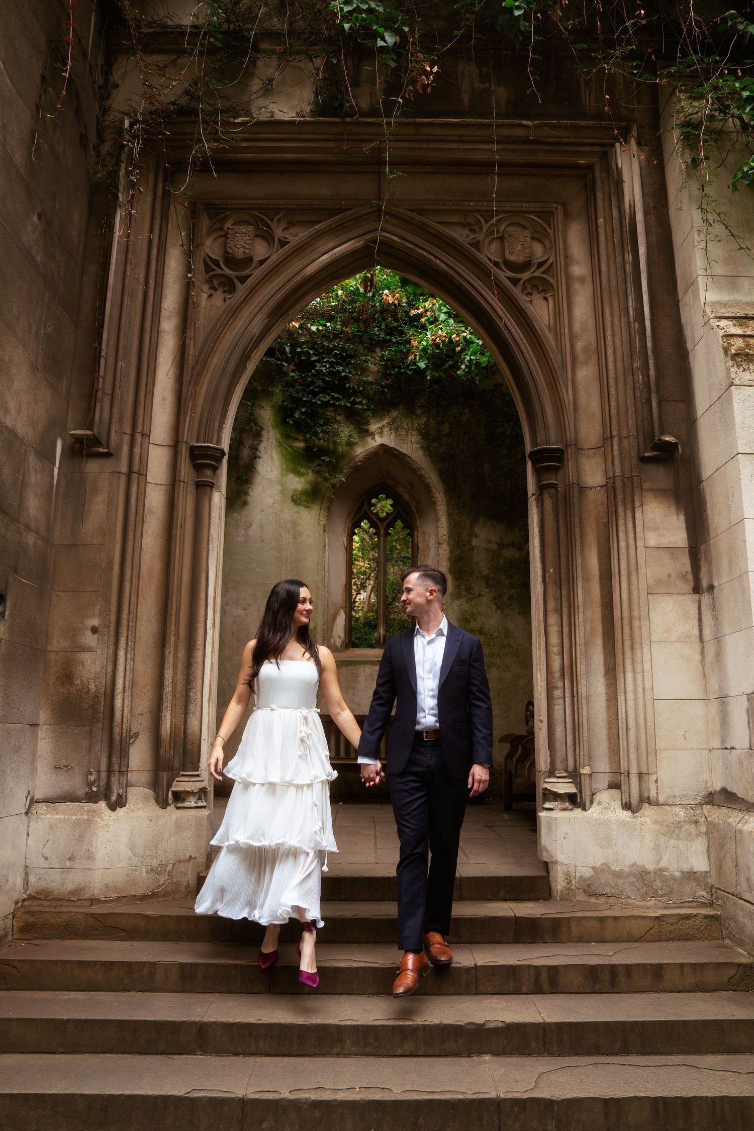 Couple in a white tiered dress and navy suit walk hand in hand down stone steps in ivy-covered church ruins.
