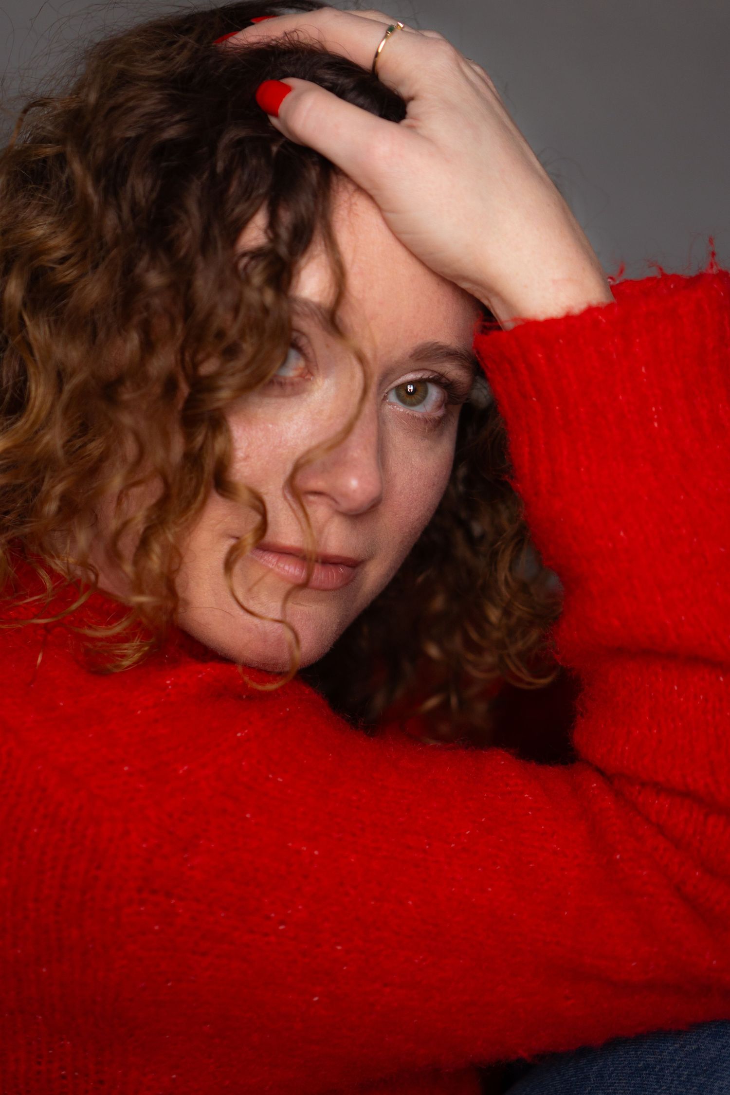 Woman with curly auburn hair and red nail polish rests her hand on her head, wearing a bright red knit sweater against a gray backdrop.