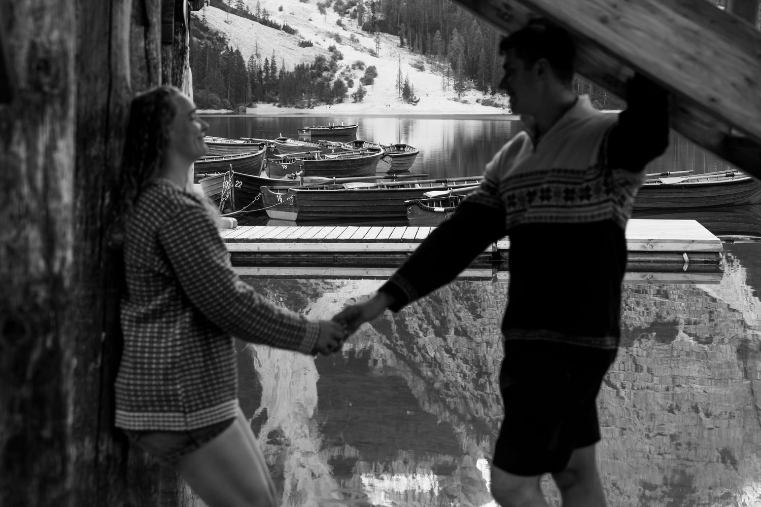Black and white photo of a couple holding hands beside a wooden structure with boats docked on a calm mountain lake behind them.