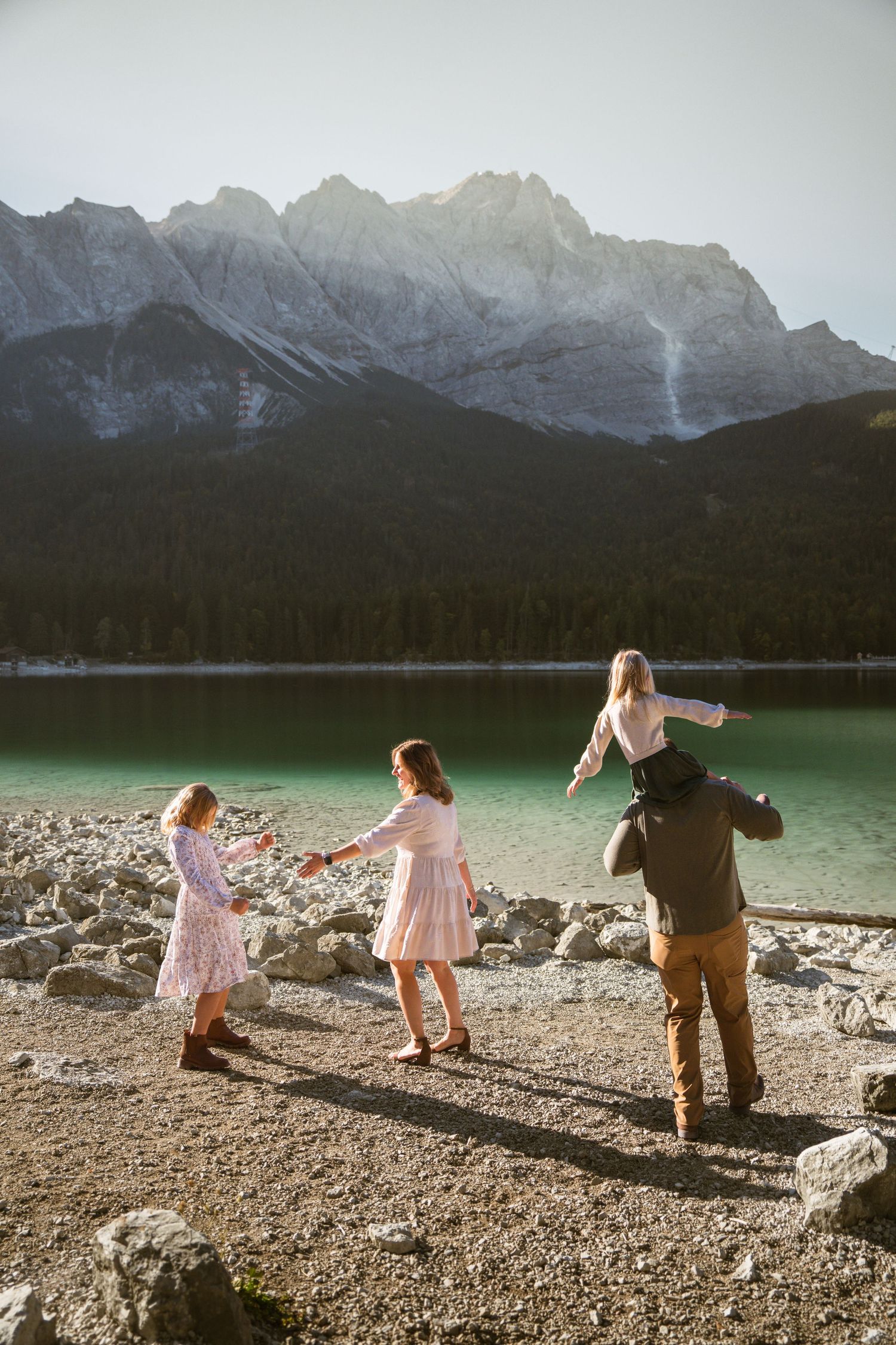 Family of five plays beside a turquoise mountain lake, with a child on dad's shoulders and two girls twirling in dresses.