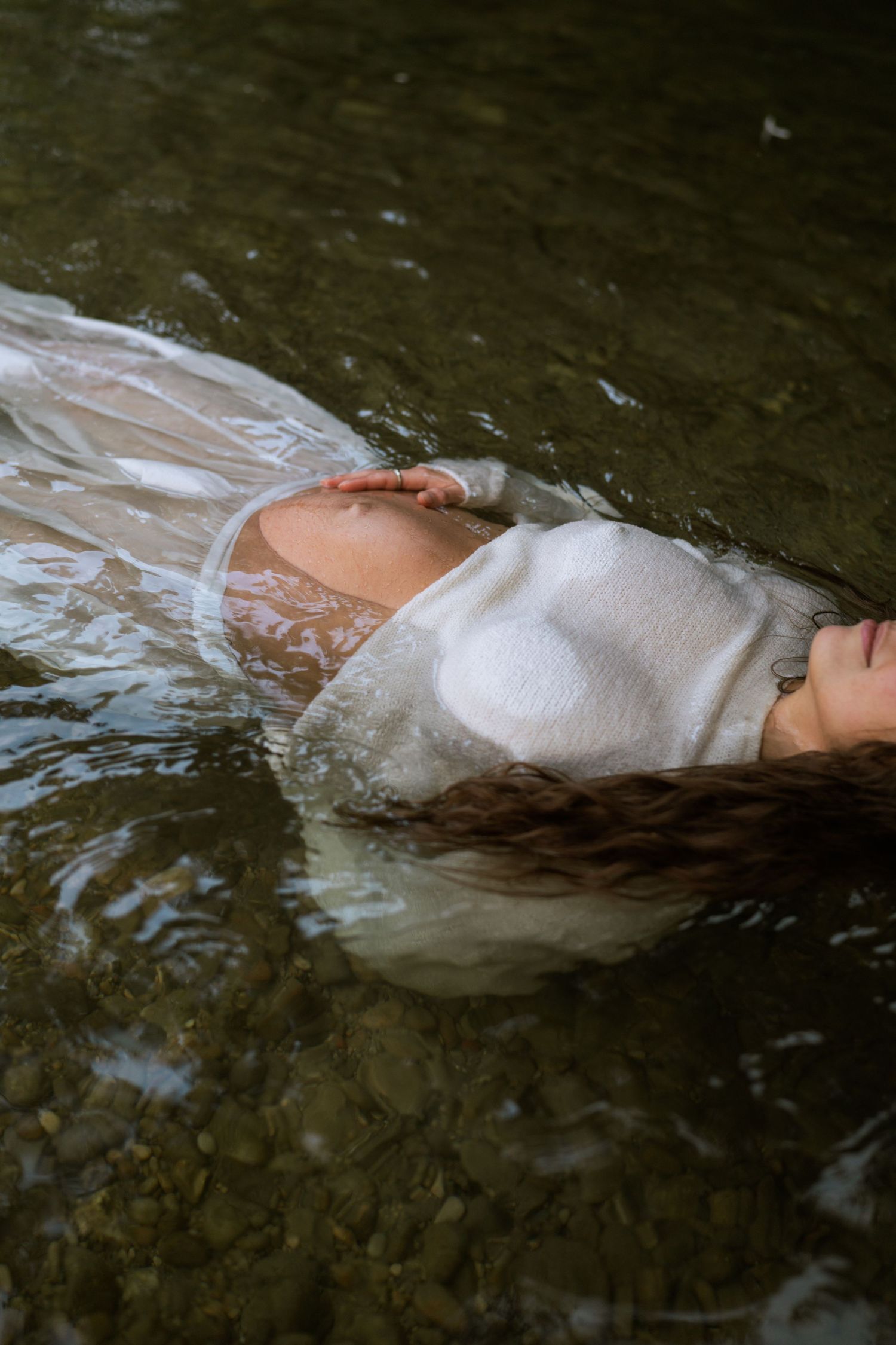 Pregnant woman in a white top floats on her back in clear shallow water, her belly and long hair resting on the surface.