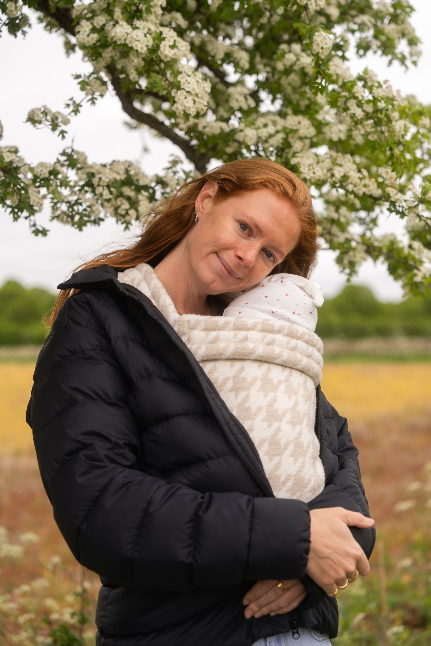 Smiling red-haired woman in a black puffer jacket holding a baby wrapped in a houndstooth carrier beneath a blossoming tree.
