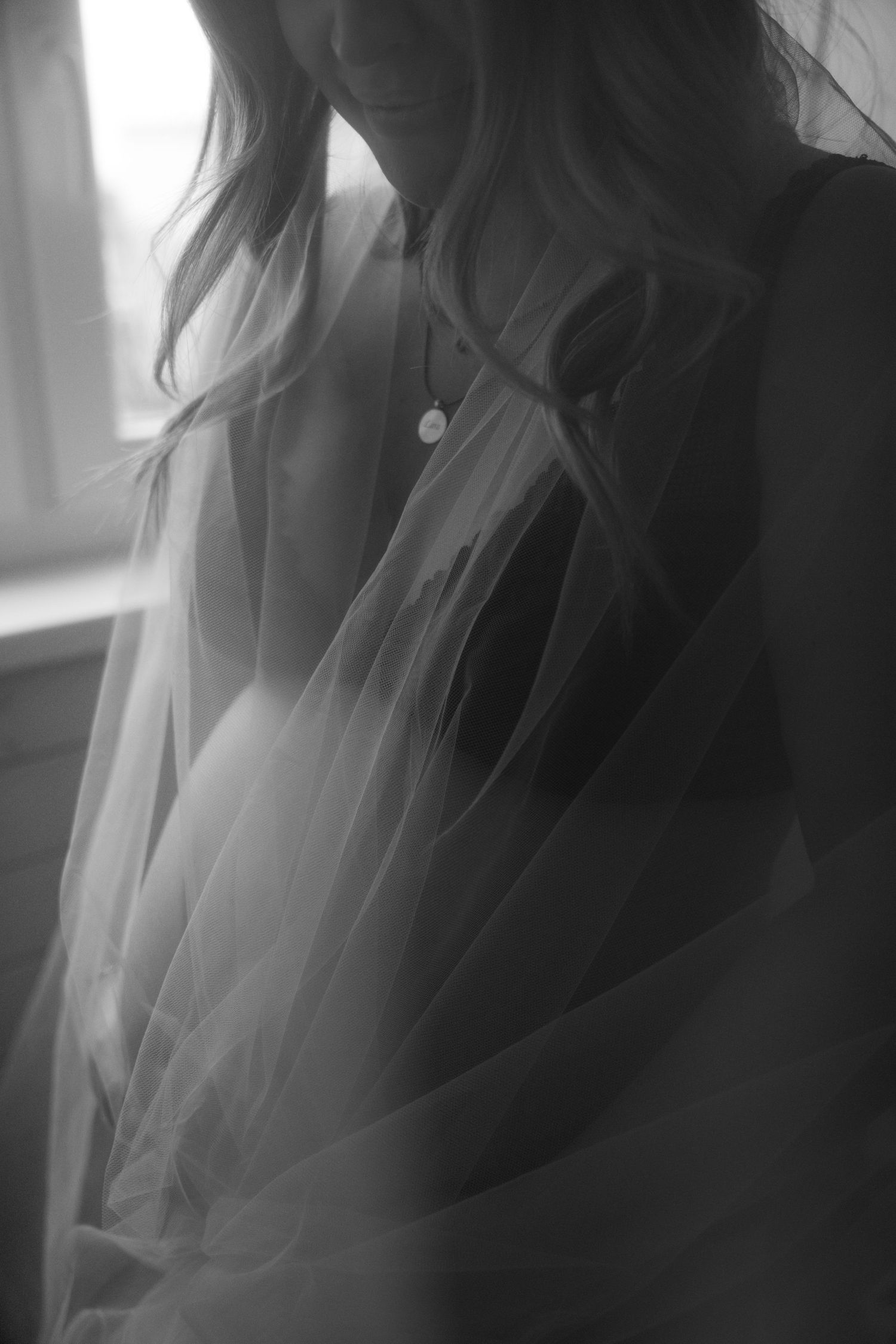 Black and white maternity portrait of a woman with long hair and a pendant necklace, wrapped in sheer tulle beside a softly lit window.