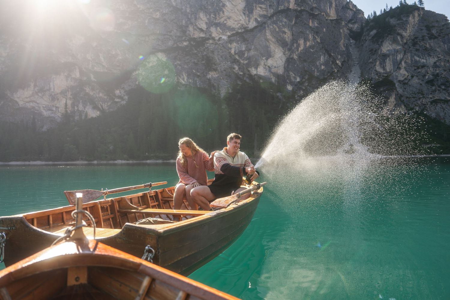 Two people sit in a wooden rowboat on turquoise alpine lake water as a spray of water splashes beside them, with rocky mountains behind.