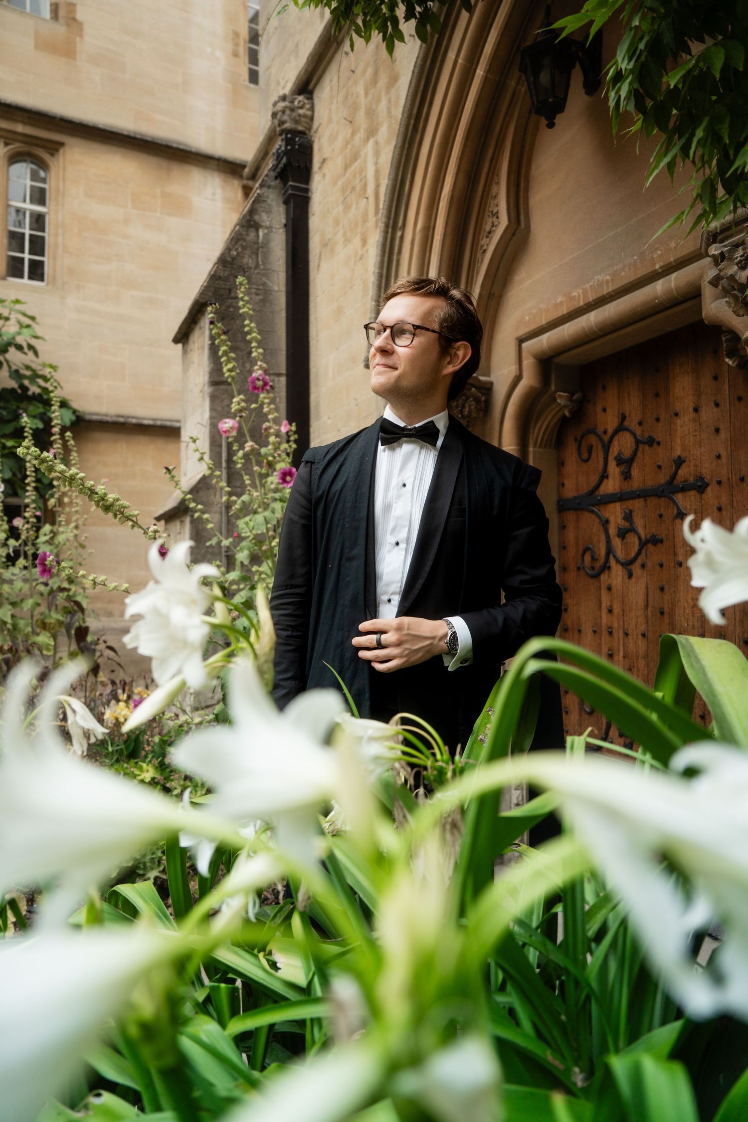 Man in glasses, black bow tie and dark jacket stands beside a stone doorway, surrounded by white lilies and green foliage.