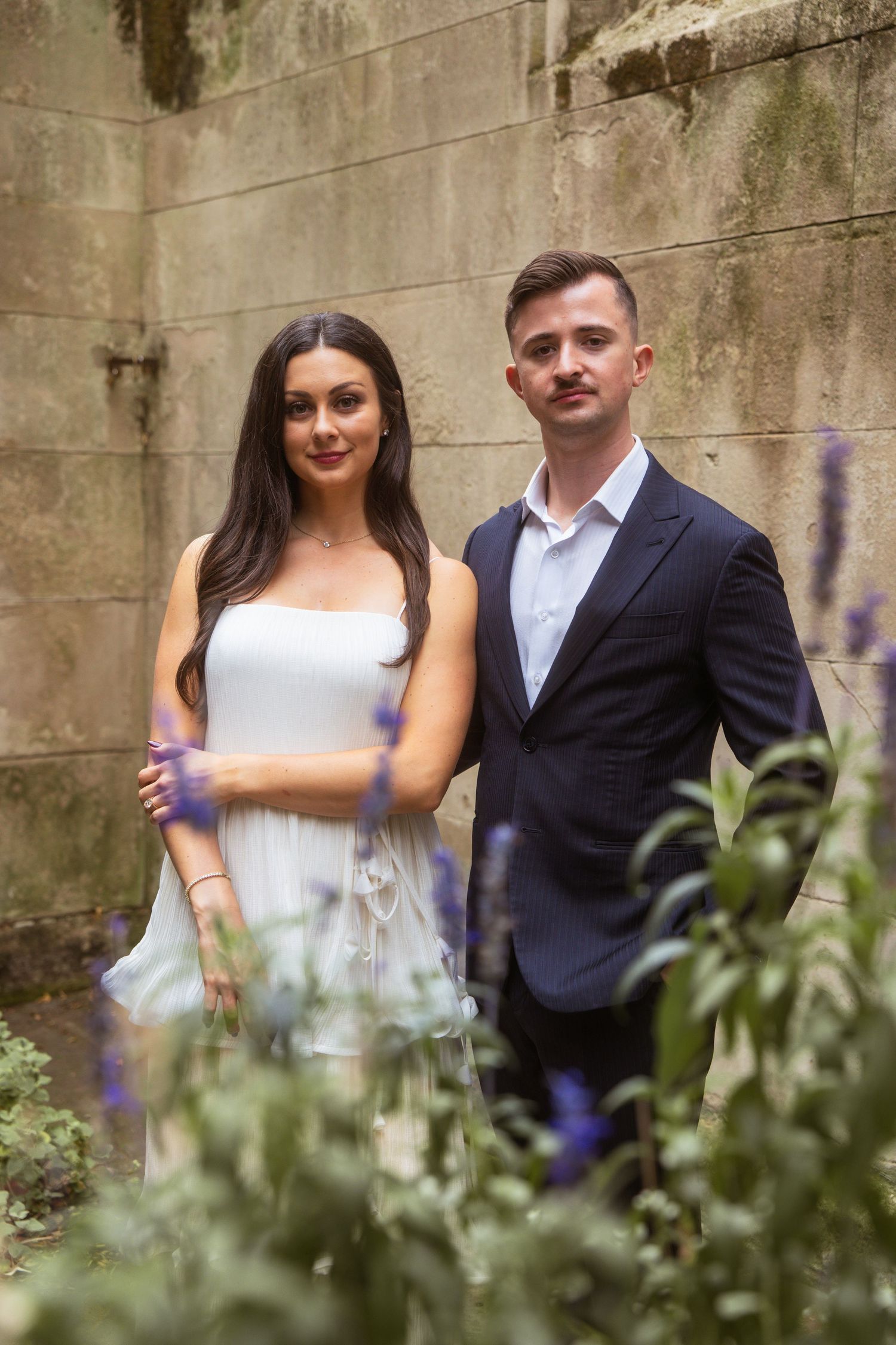 Engagement portrait of a smiling couple in white dress and navy suit among lavender in a mossy stone courtyard.