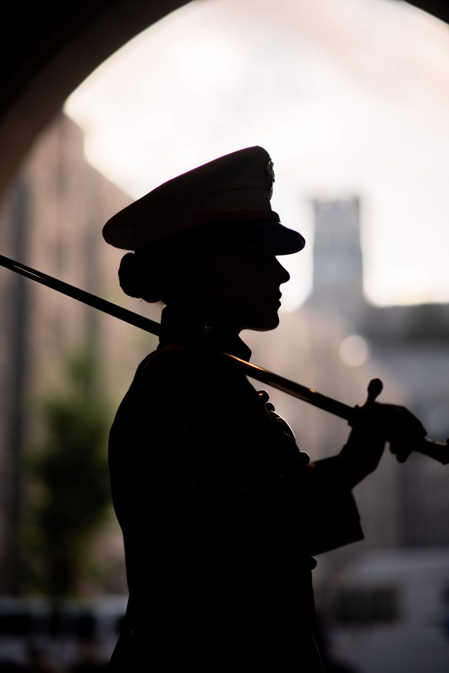 Silhouette of a uniformed soldier in a peaked cap carrying a sword on the shoulder beneath a stone archway.