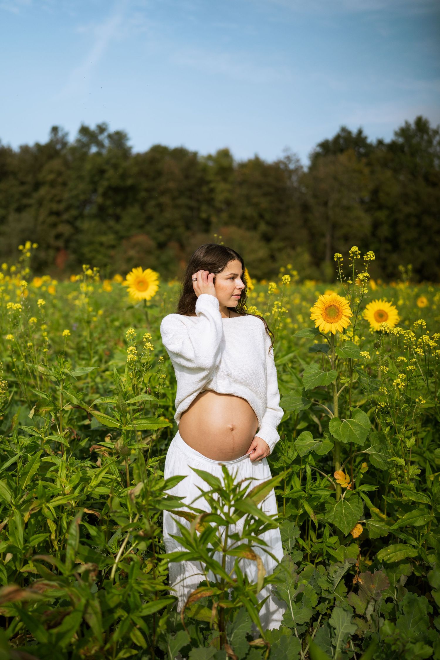 Pregnant woman in white stands among blooming sunflowers in a sunny field with a tree line behind her.