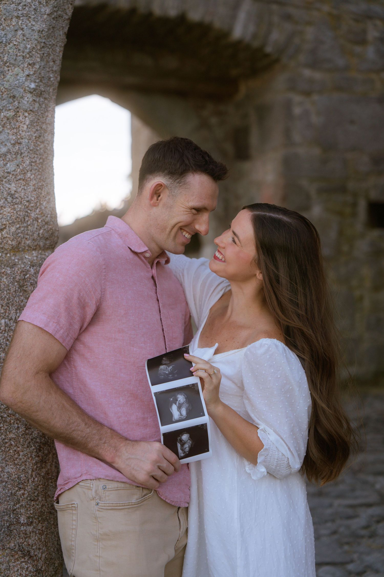 Smiling couple embracing against a stone archway while holding ultrasound photos, a pregnancy announcement portrait.