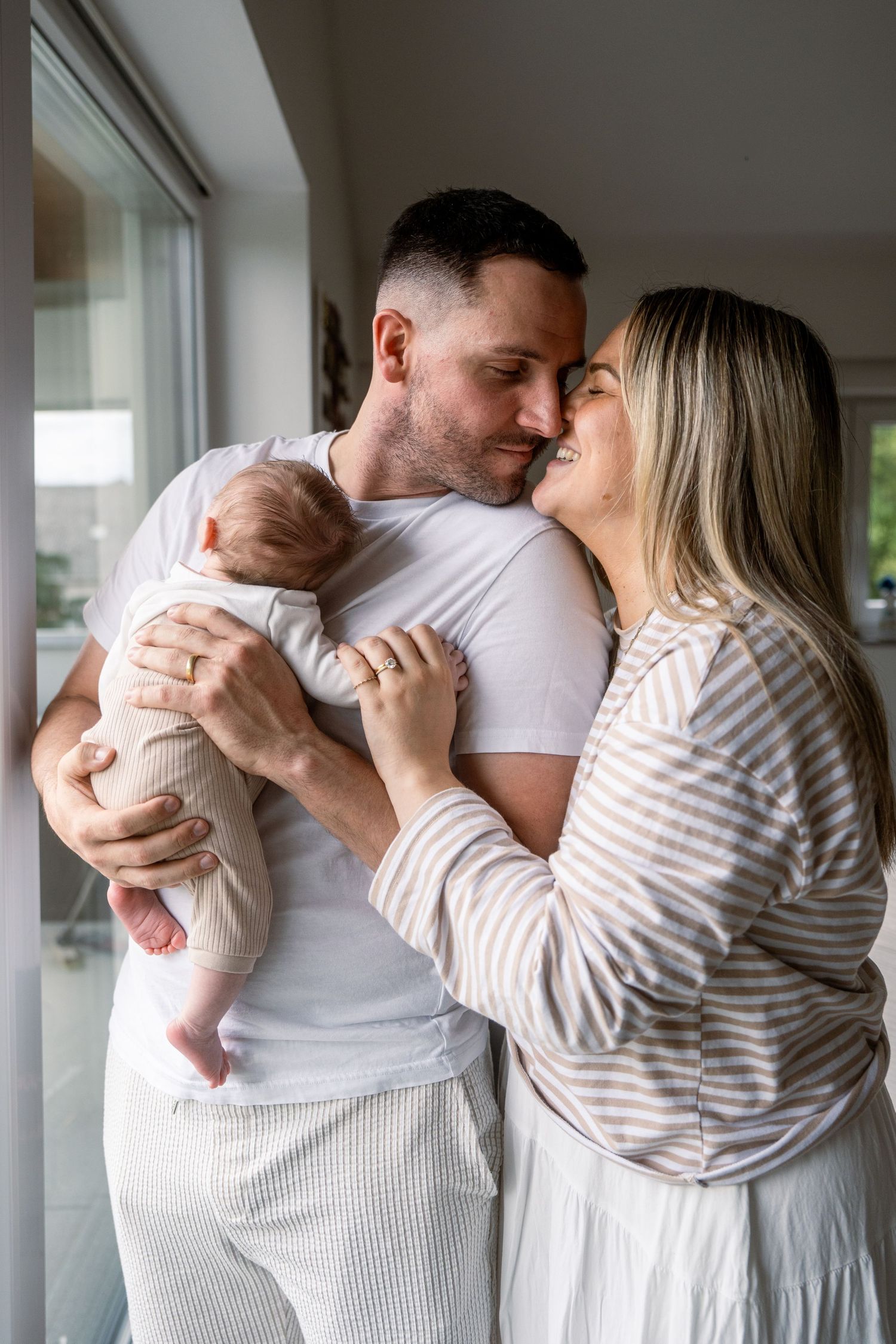 Smiling couple touching foreheads while gently holding their sleeping newborn baby beside a bright window.