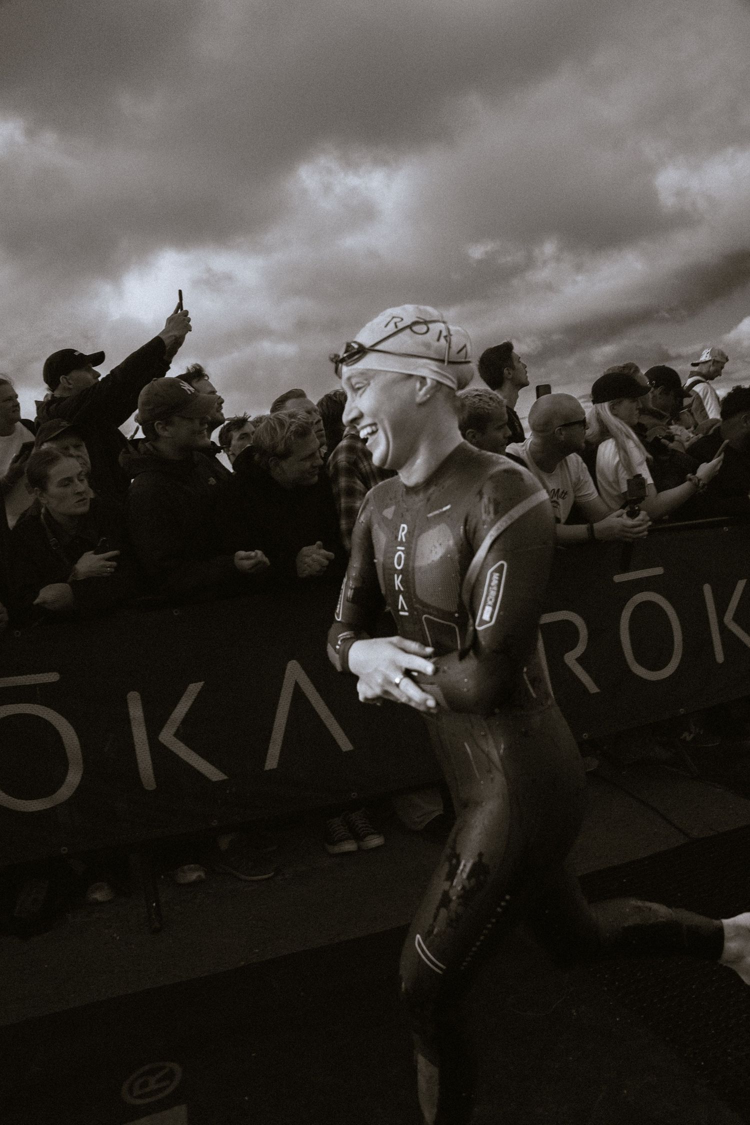 Smiling triathlete in a wetsuit and swim cap runs past cheering spectators behind barriers under a dramatic cloudy sky in black and white.