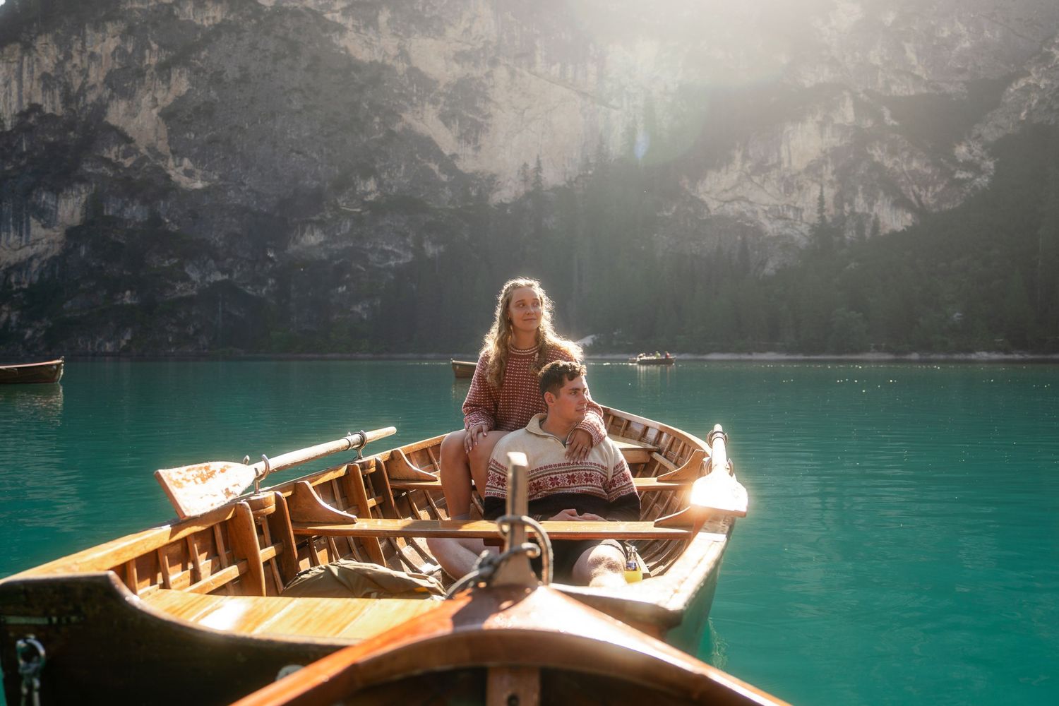 Two people relax in a wooden rowboat on calm turquoise alpine lake water beneath rocky mountain cliffs in warm golden sunlight.