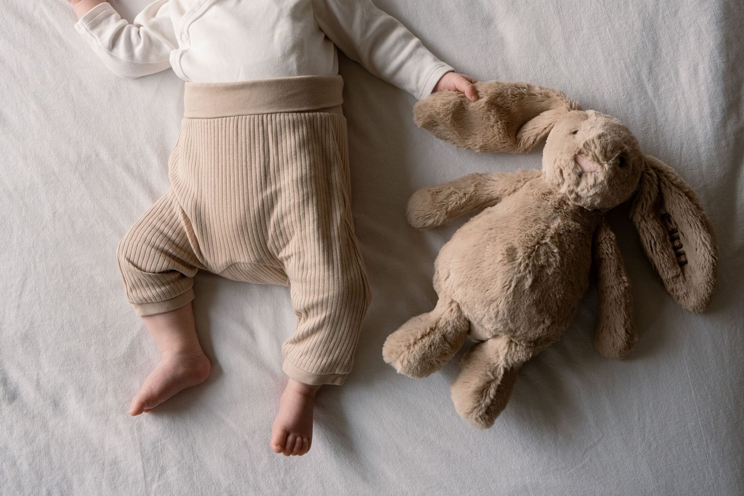 Baby in cream ribbed pants and white top lying barefoot on soft bedding beside a plush beige stuffed bunny toy.