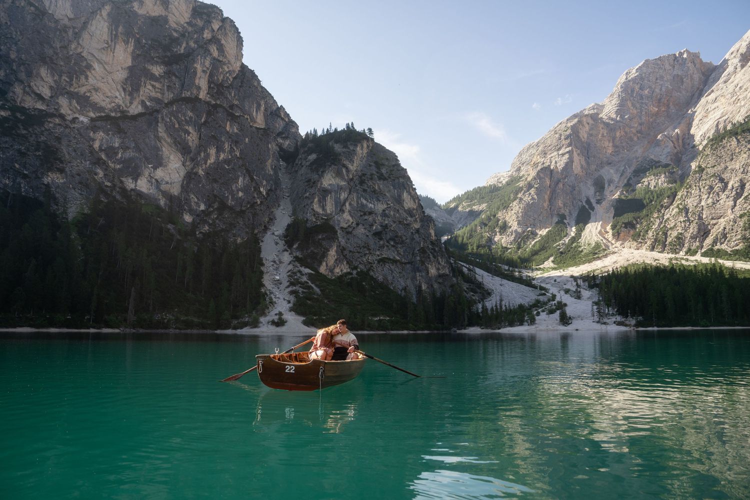 Wooden rowboat with two people glides across turquoise alpine lake, surrounded by towering rocky Dolomites mountains and evergreen forest.