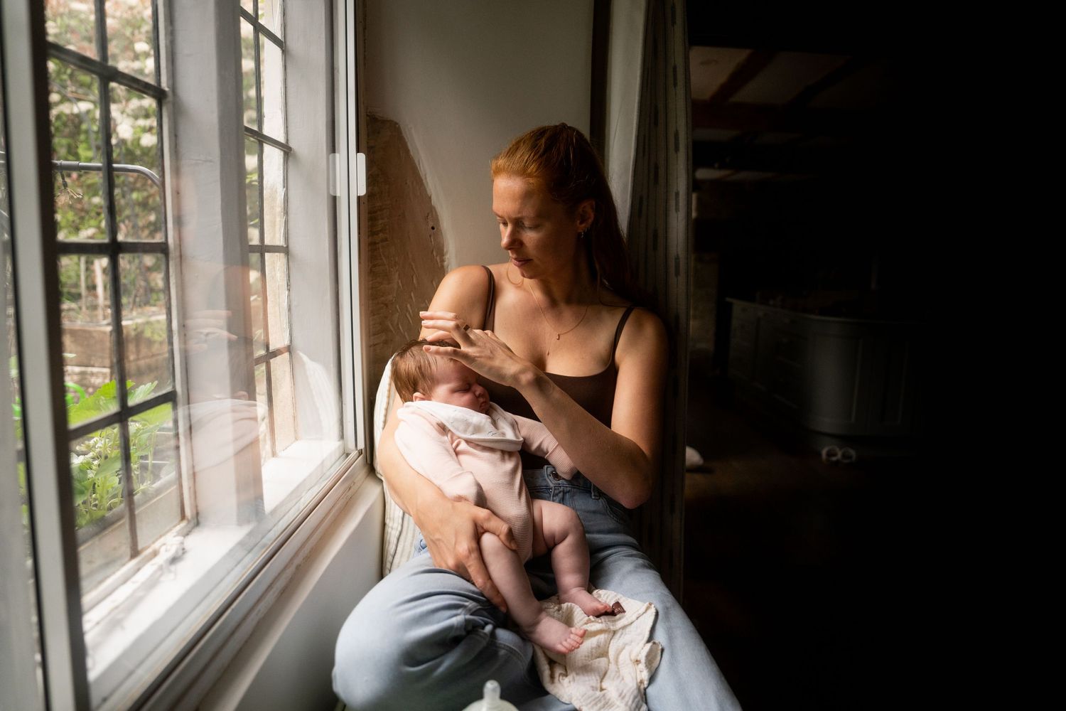 Mother sitting by a sunlit window cradling her newborn baby in a quiet, softly lit room with green trees visible outside.