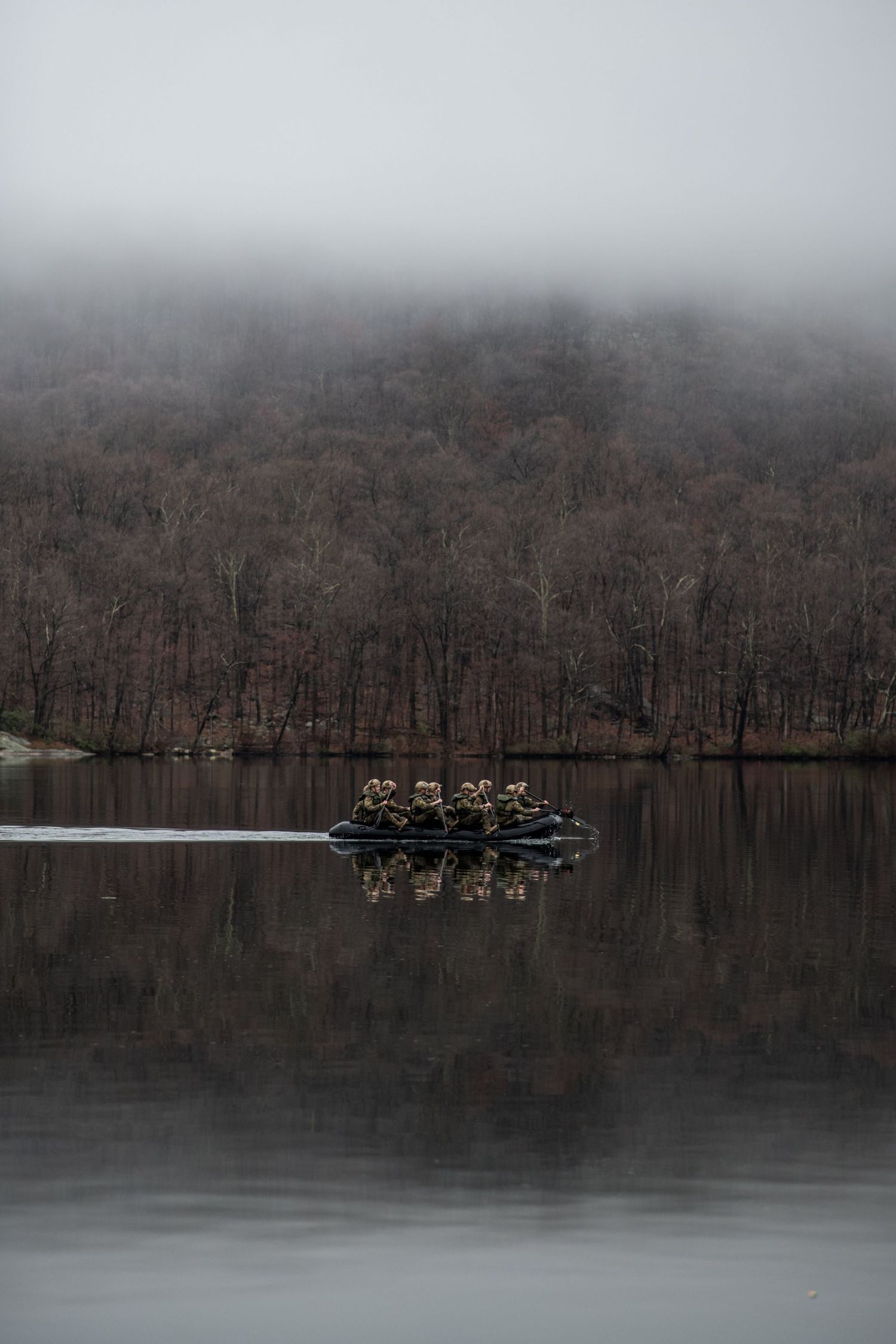 A small inflatable raft carrying a team glides across a still, misty lake beneath a fog-covered wooded hillside.