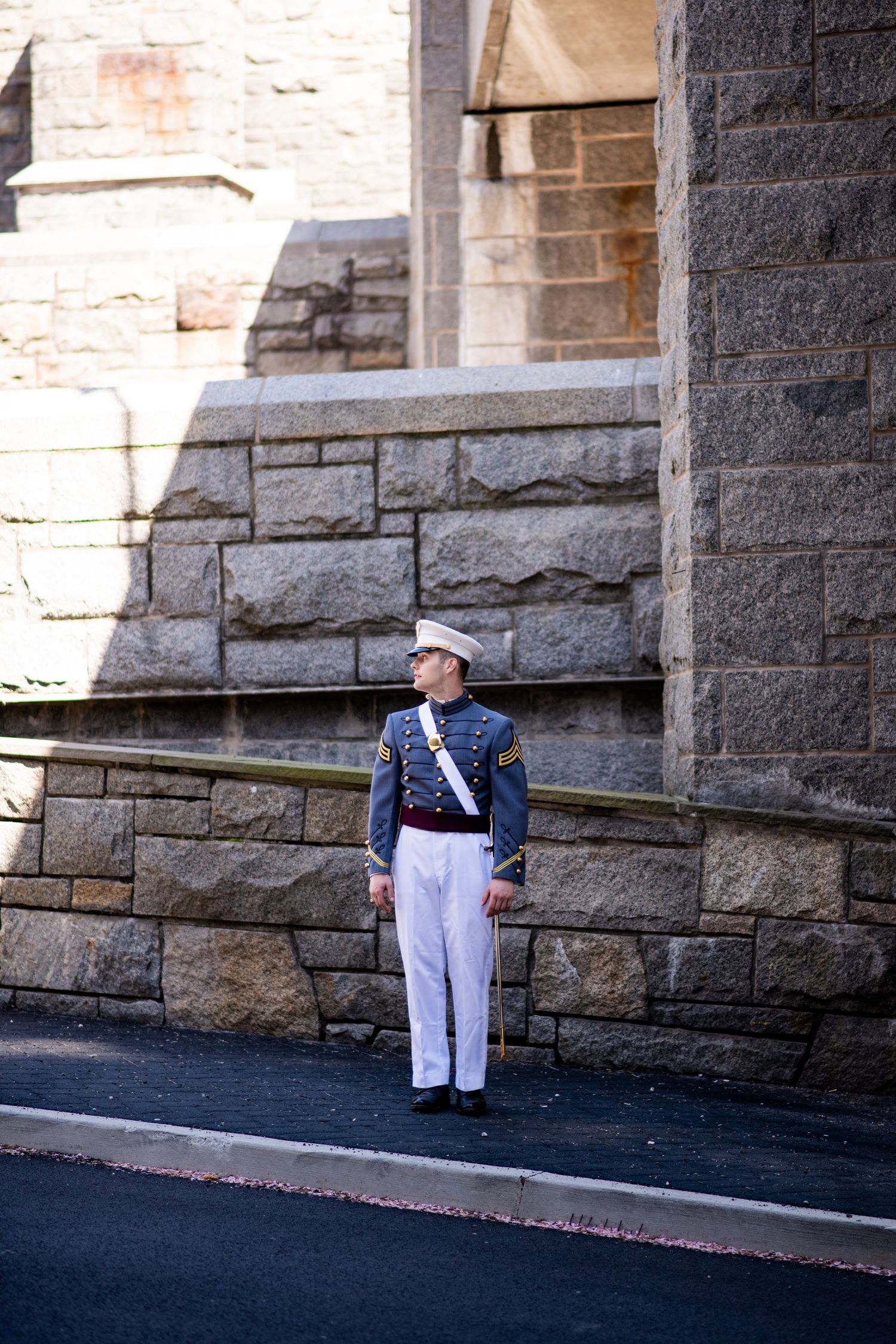 A uniformed cadet in gray jacket, white trousers and white cap stands beside a tall stone wall, looking to the side.