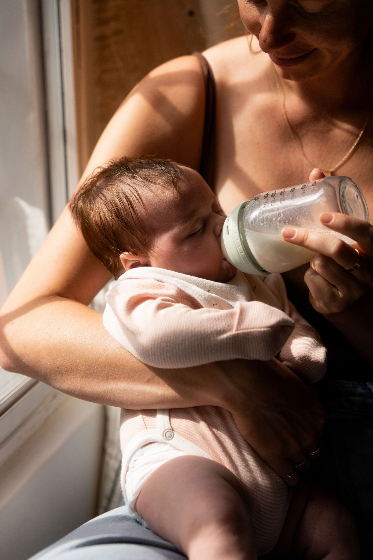 A smiling mother cradles her sleepy newborn in soft sunlight by a window while feeding the baby milk from a glass bottle.