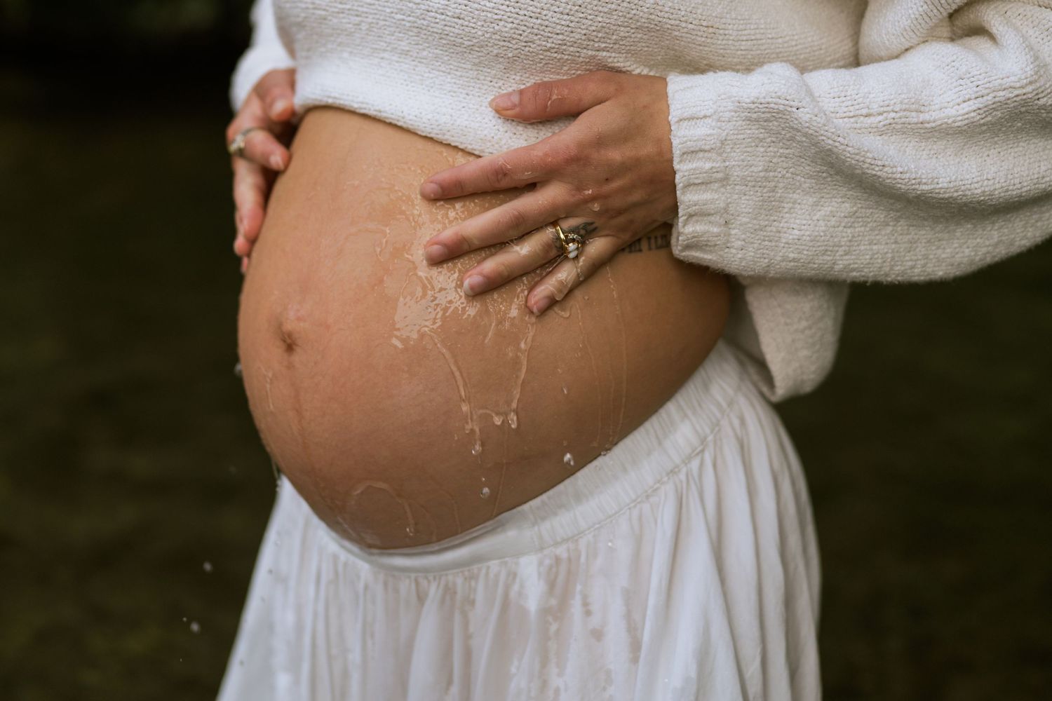 Close-up of a pregnant woman's bare belly with golden paint accents as she rests her hands on it in a white sweater and skirt.