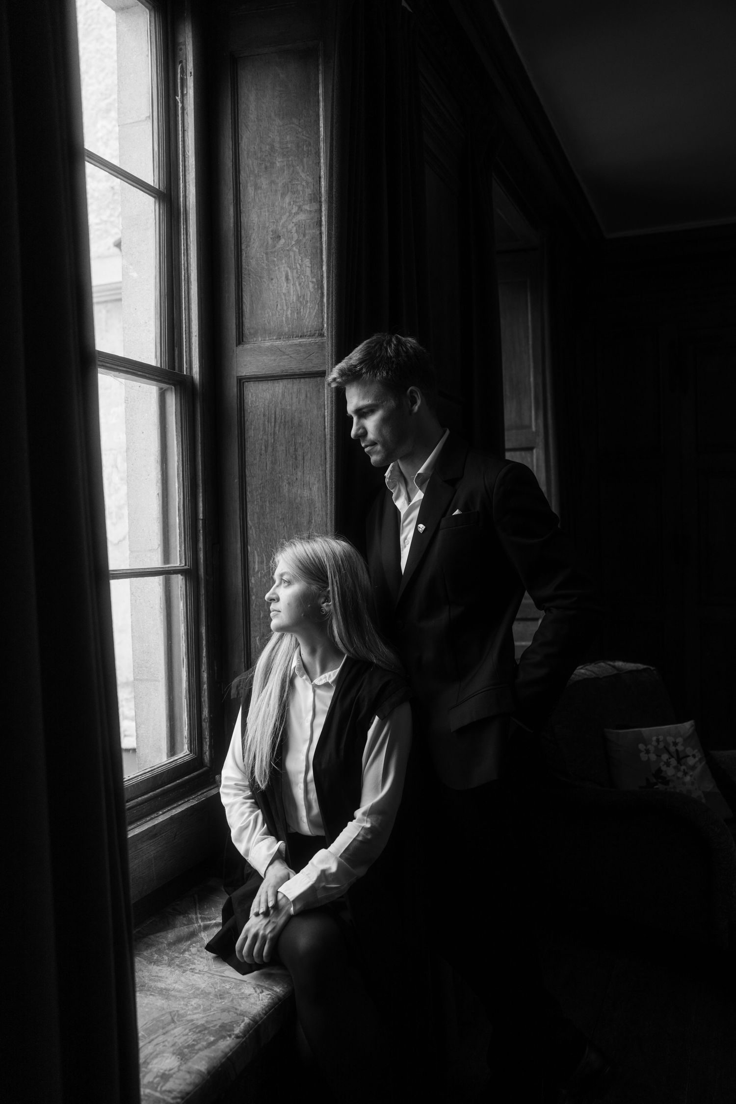 Black and white portrait of a man in a blazer standing behind a seated woman as both gaze out a tall window in a dimly lit paneled room.