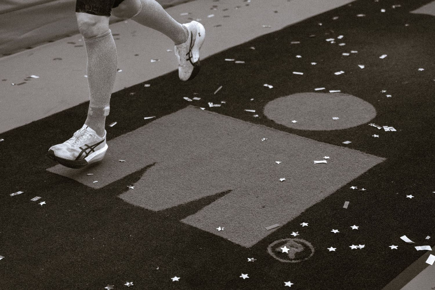 Runner in white athletic shoes steps over a gray timing mat on a black road covered in scattered confetti during a race.