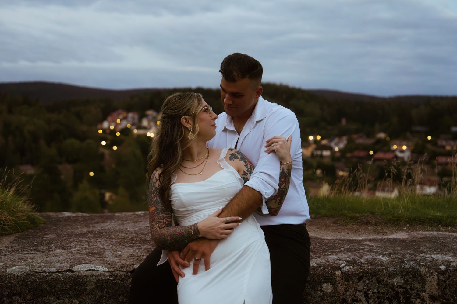 Engaged couple embracing at dusk on a stone wall overlooking a hillside town with glowing lights.