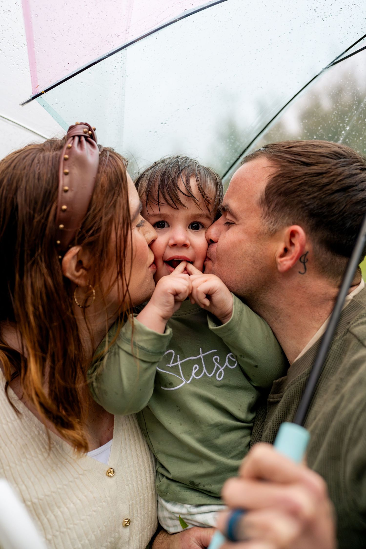 A couple kisses their smiling toddler on both cheeks while sheltering together under a clear umbrella on a rainy day.
