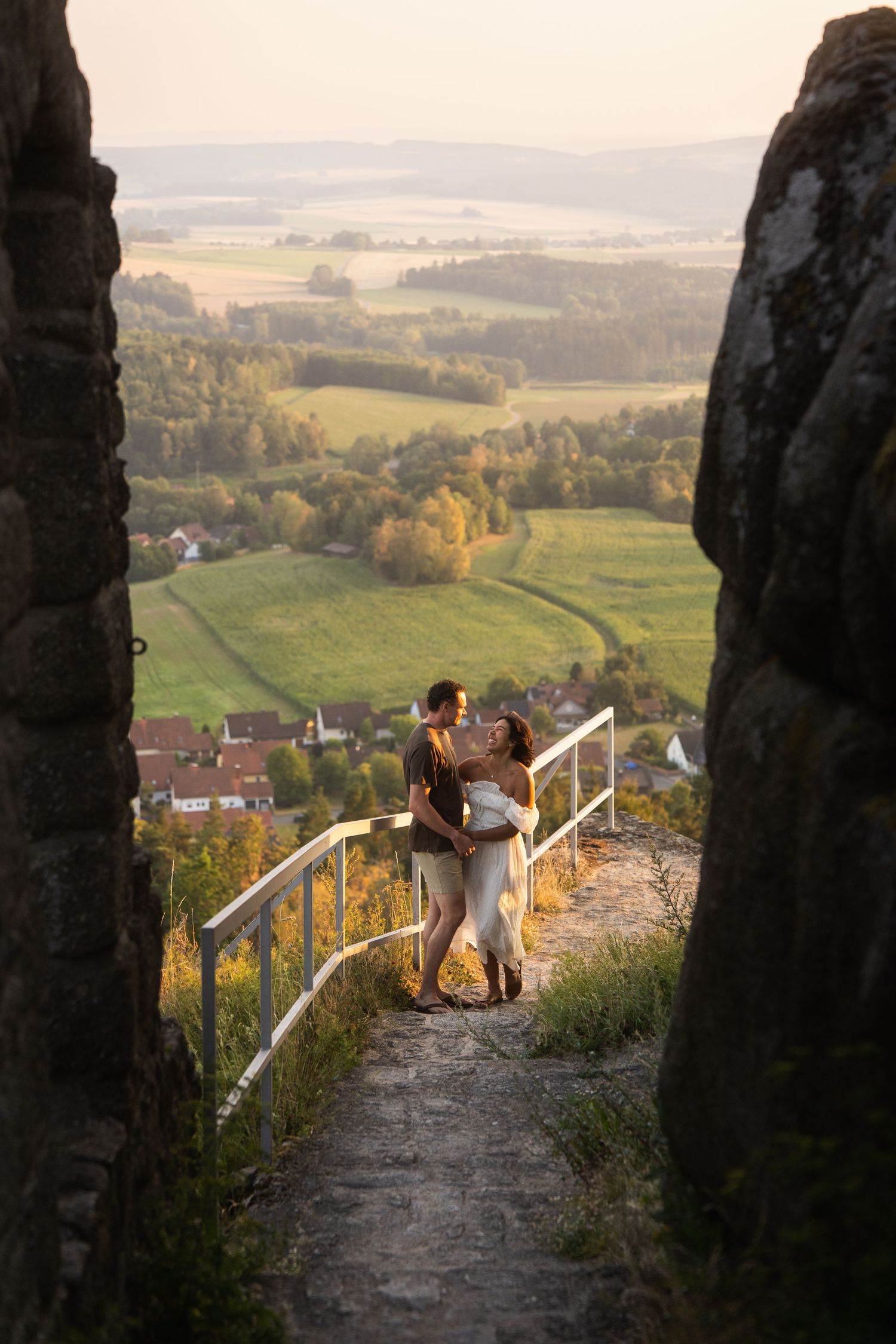 Couple embracing on a stone path between rock walls at sunset, overlooking green valley meadows, forests and village rooftops.