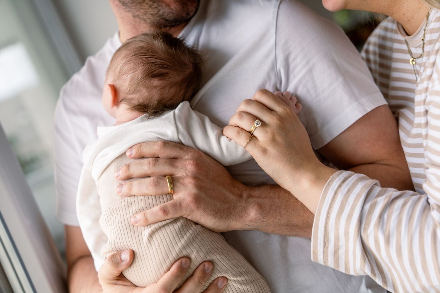 Parents with wedding rings gently cradle a sleeping newborn baby wrapped in a soft cream blanket near a bright window.