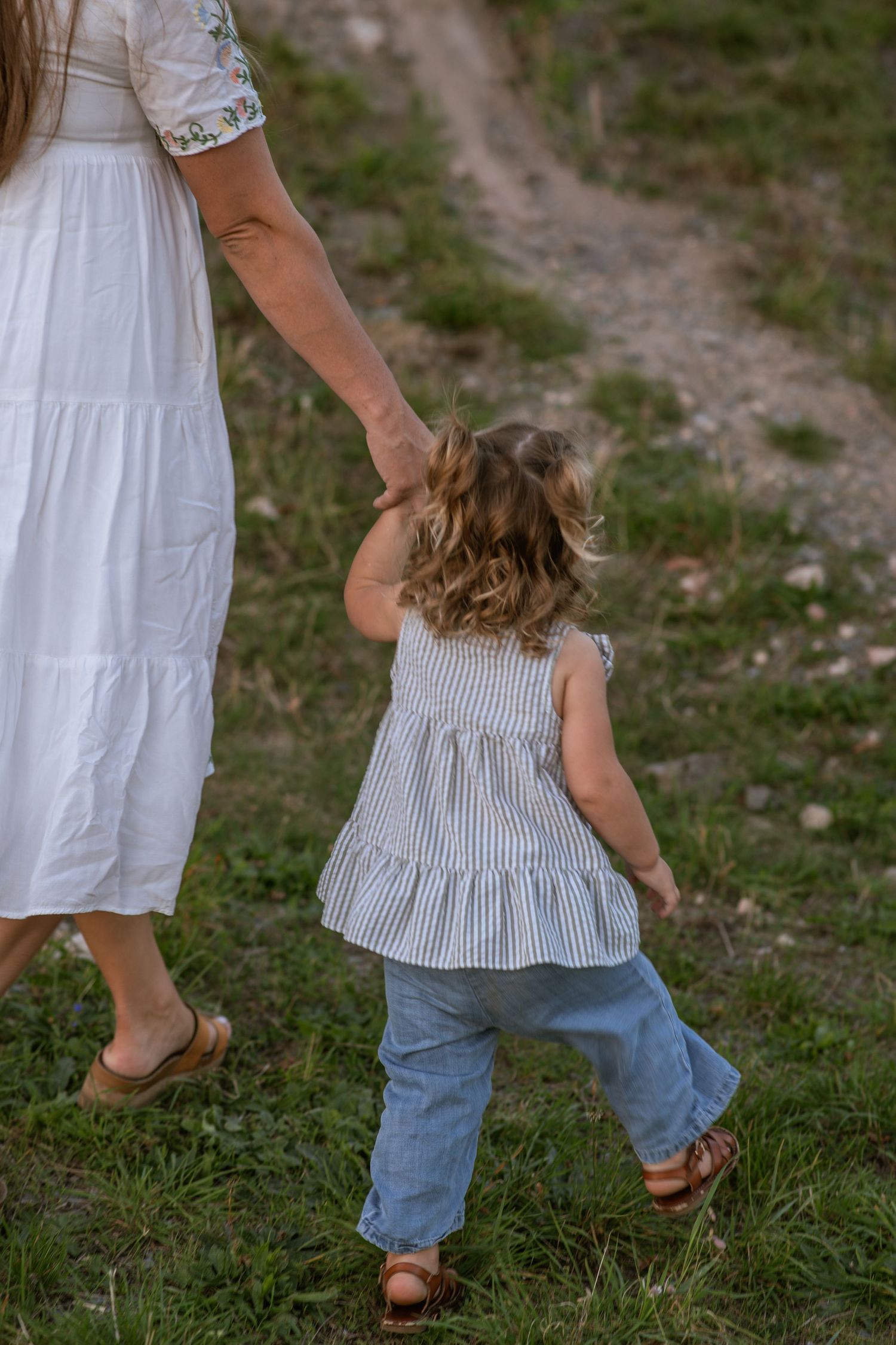 Woman in a white dress holds the hand of a toddler in a striped top and jeans while walking across a grassy hillside.