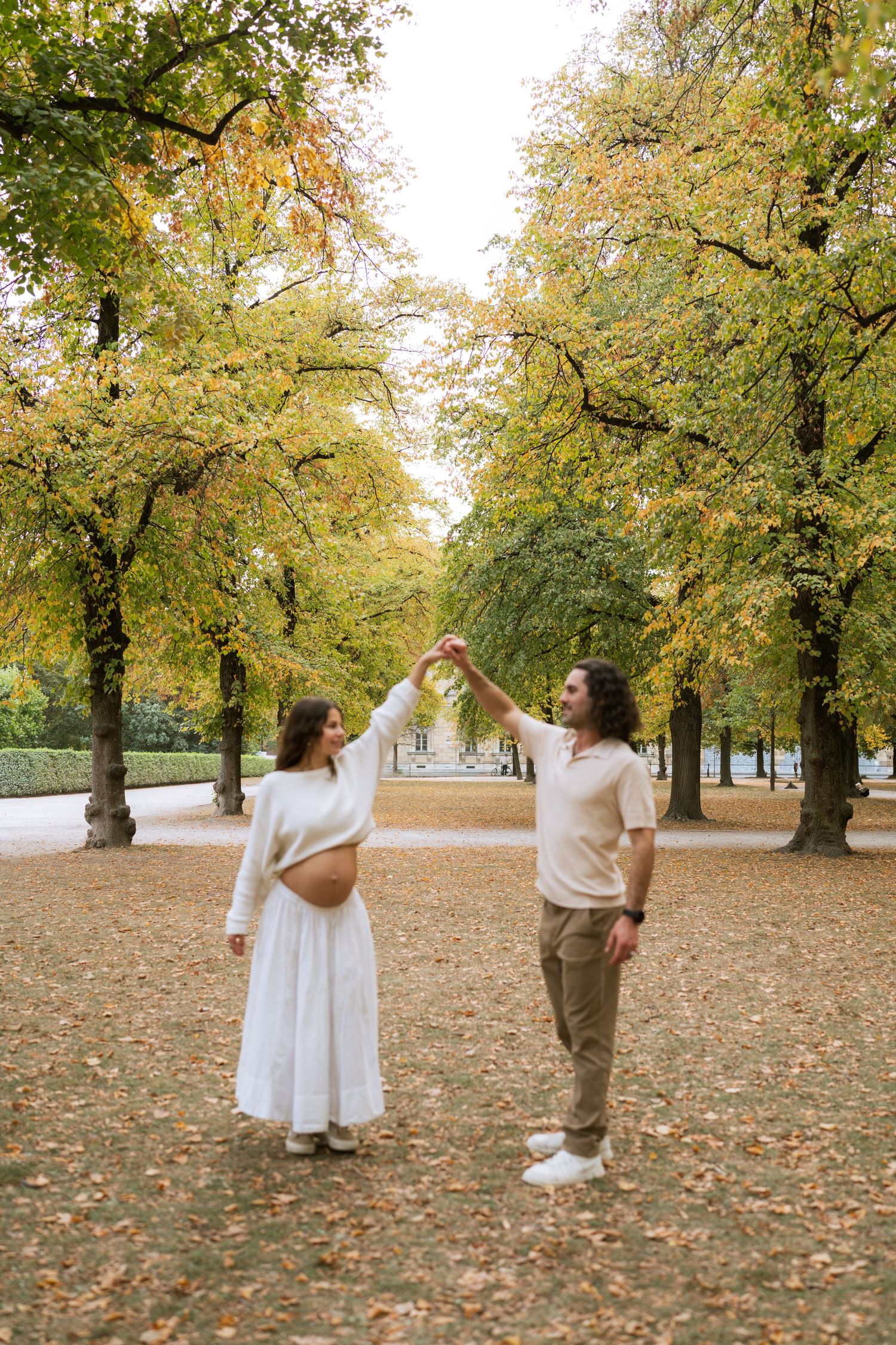 Pregnant woman in a white skirt twirls under her partner's raised hand while dancing among autumn trees in a park.