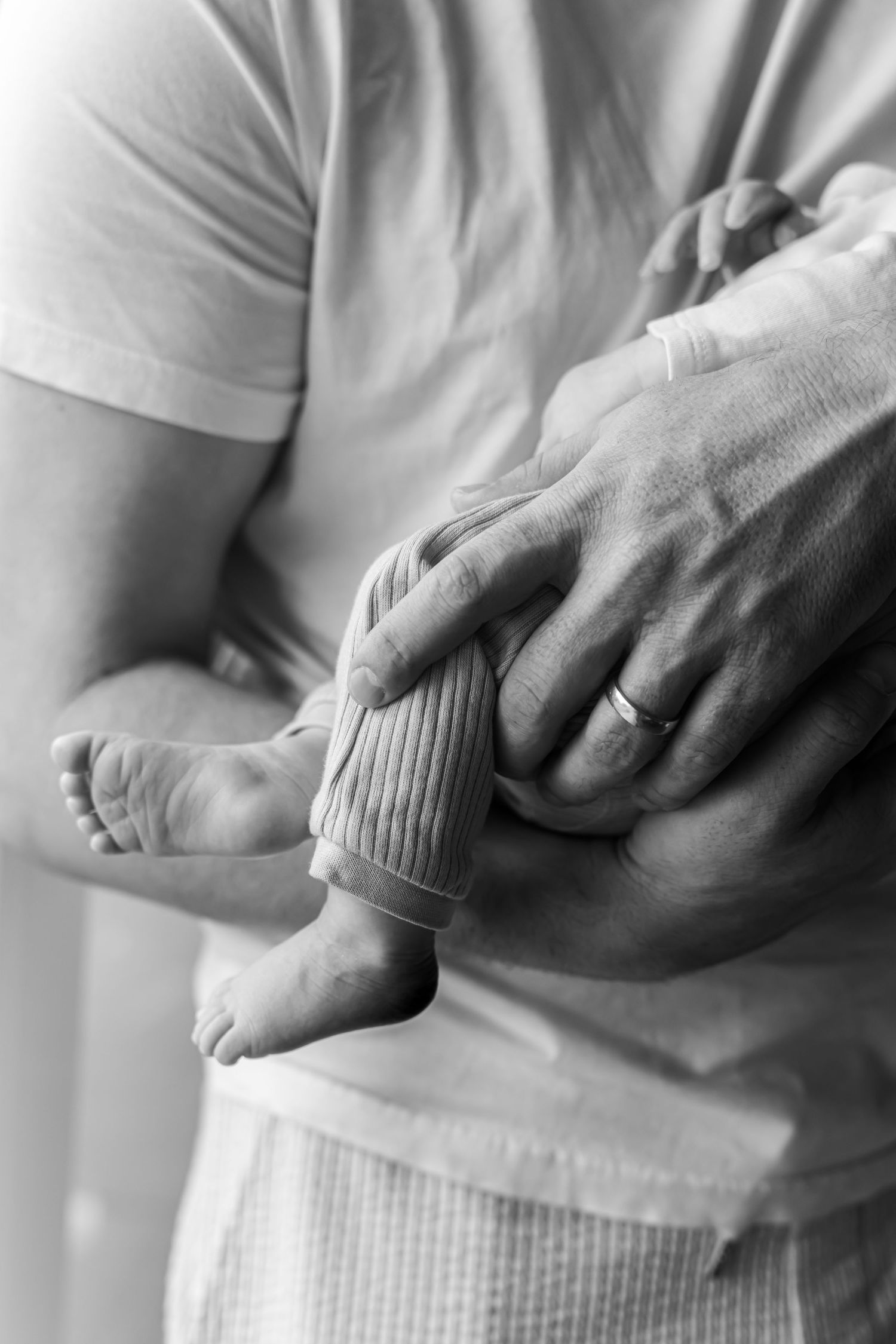 Black and white photo of a father with a wedding ring gently cradling a newborn baby's bare feet and ribbed knit outfit.