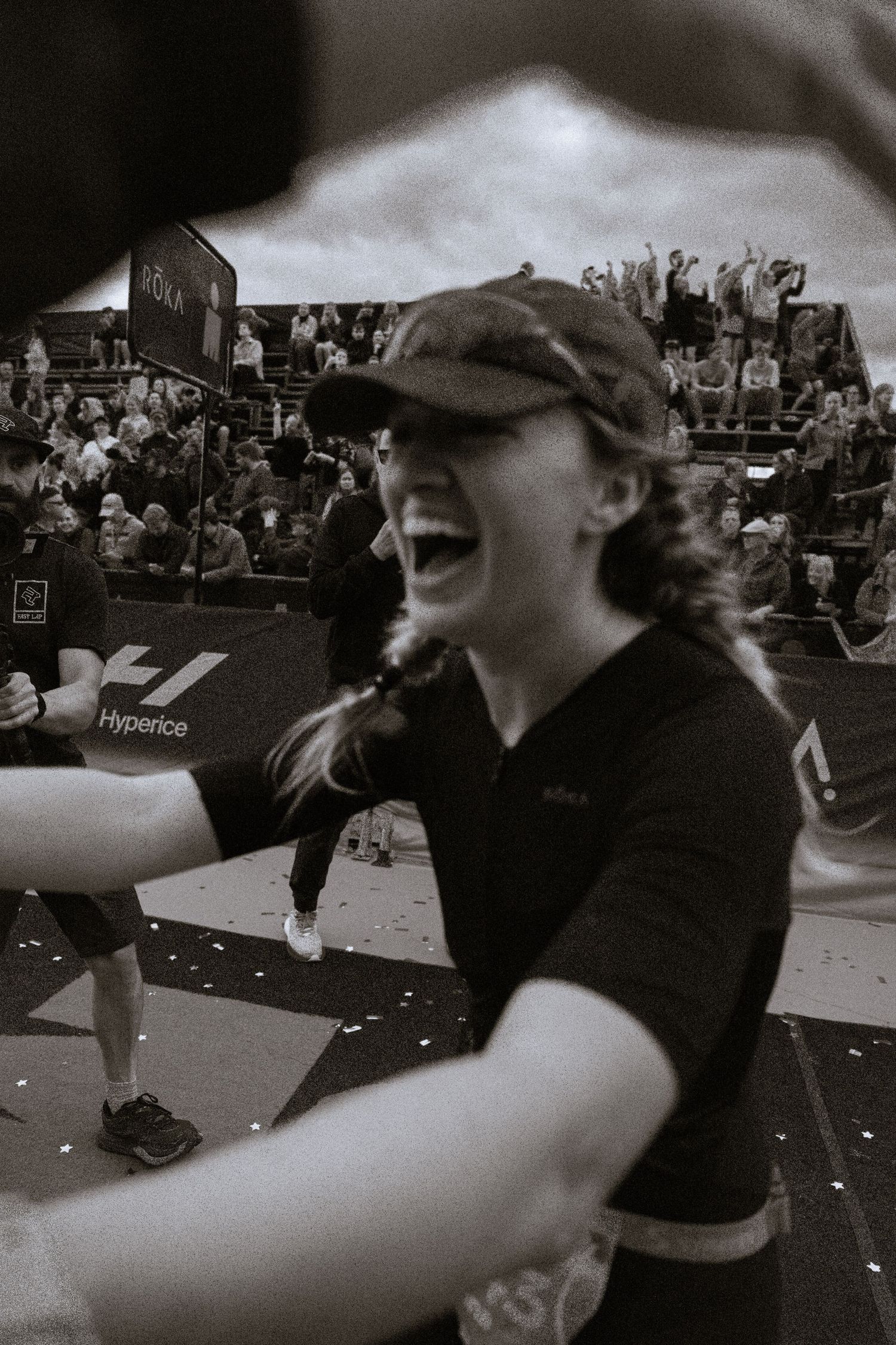Joyful runner in a cap and athletic top laughs with arms outstretched at a race finish line as spectators cheer in the grandstand behind.