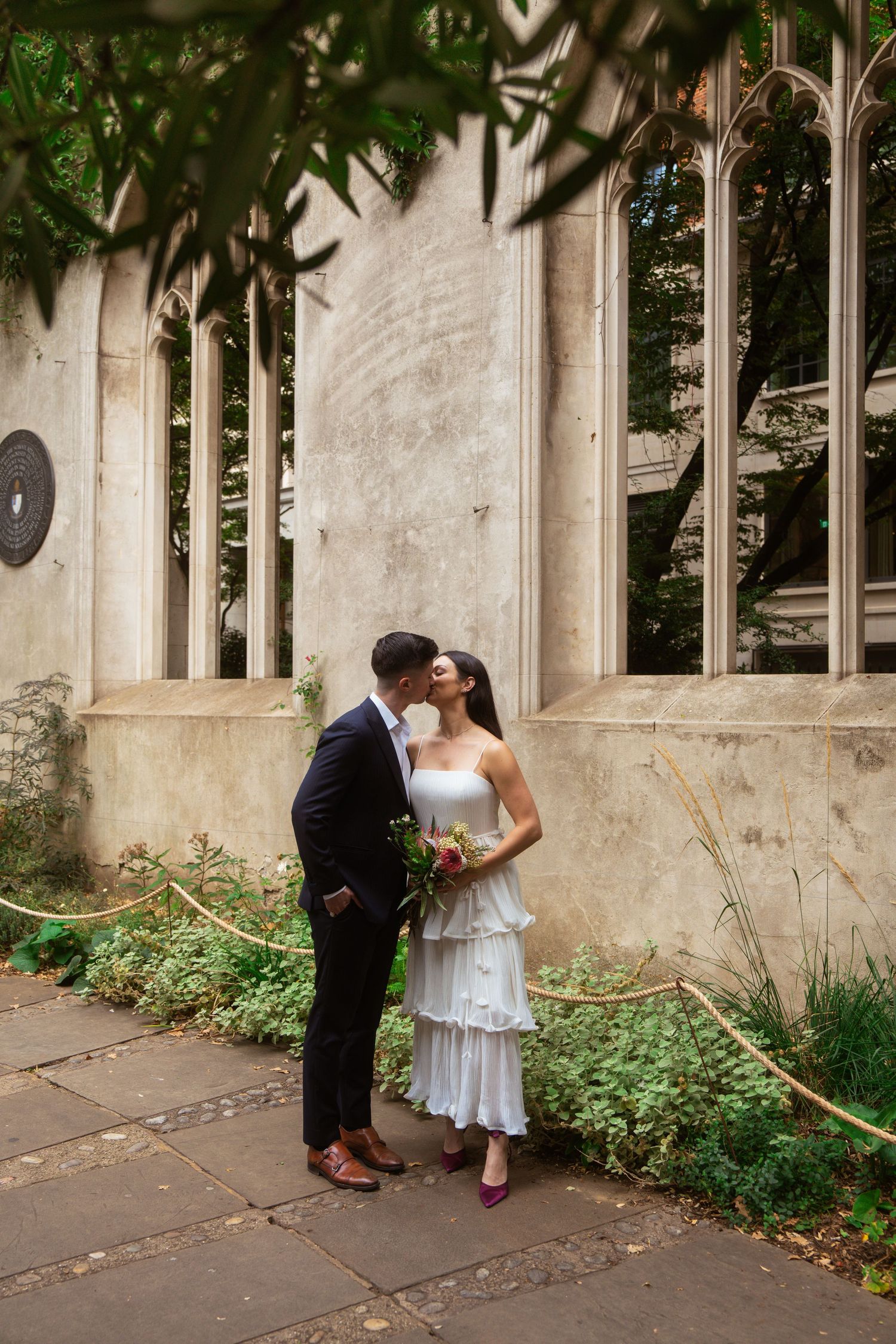 Couple kissing in a garden of historic church ruins, the bride in a white tiered dress holding a bouquet.