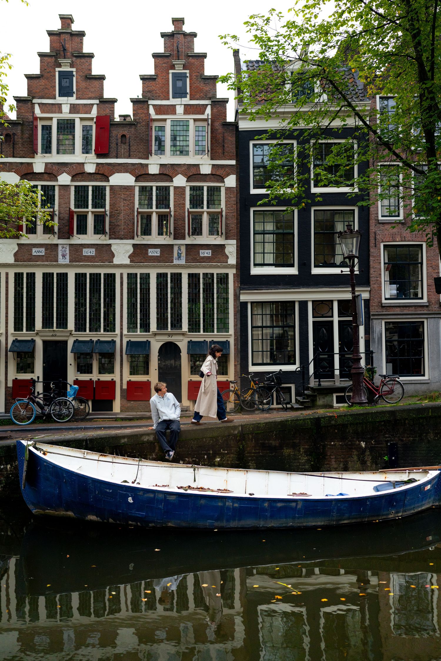Couple sitting on a canal edge beside a blue boat, with historic Dutch gabled houses in Amsterdam behind them.