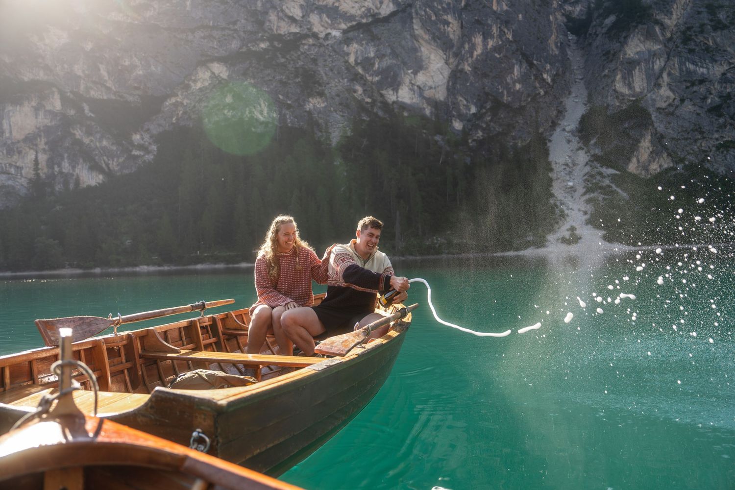 Couple relaxes in a wooden rowboat on a turquoise mountain lake with steep rocky cliffs behind them.