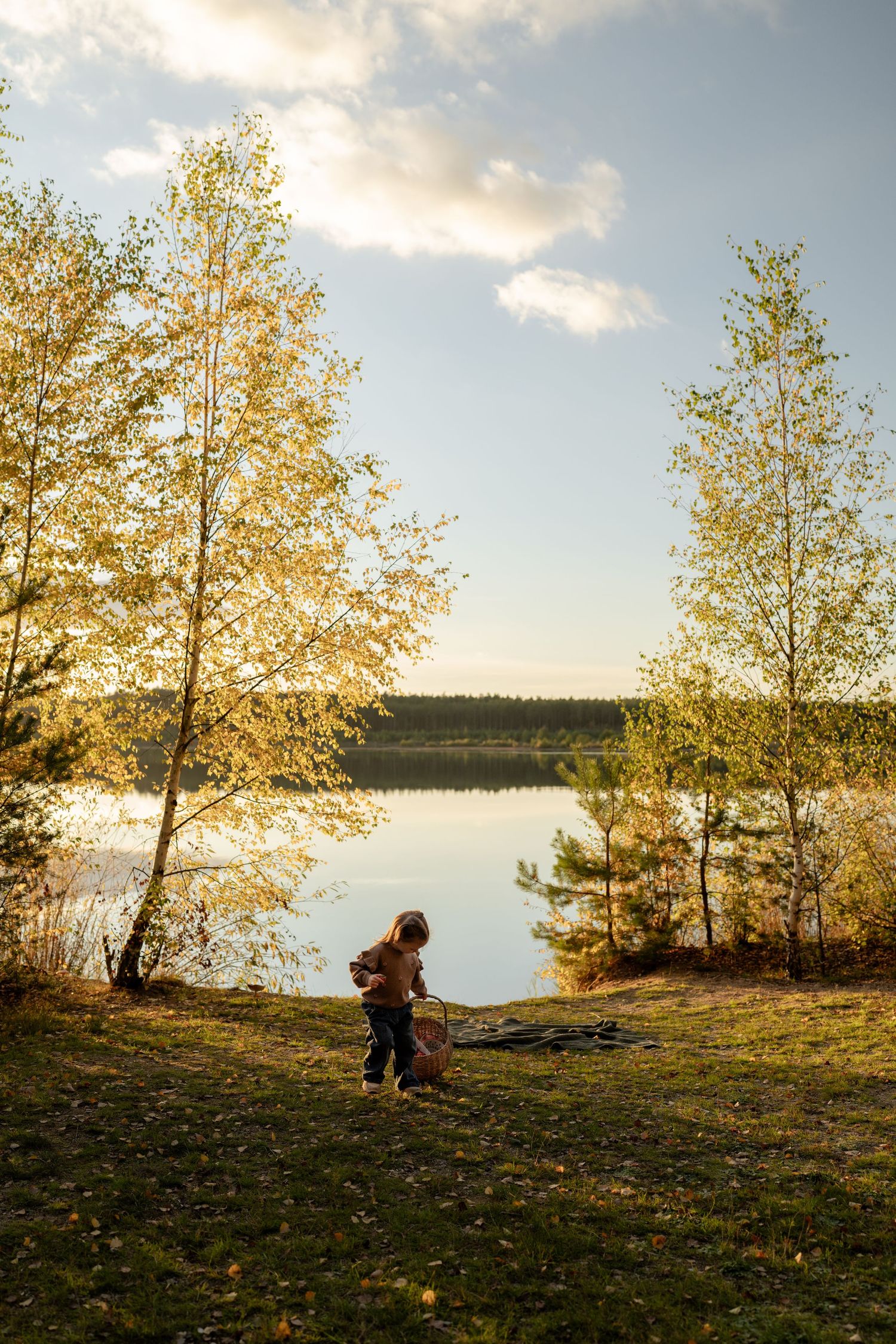 Toddler carrying a small basket explores a grassy lakeshore framed by golden birch trees in warm autumn sunlight.