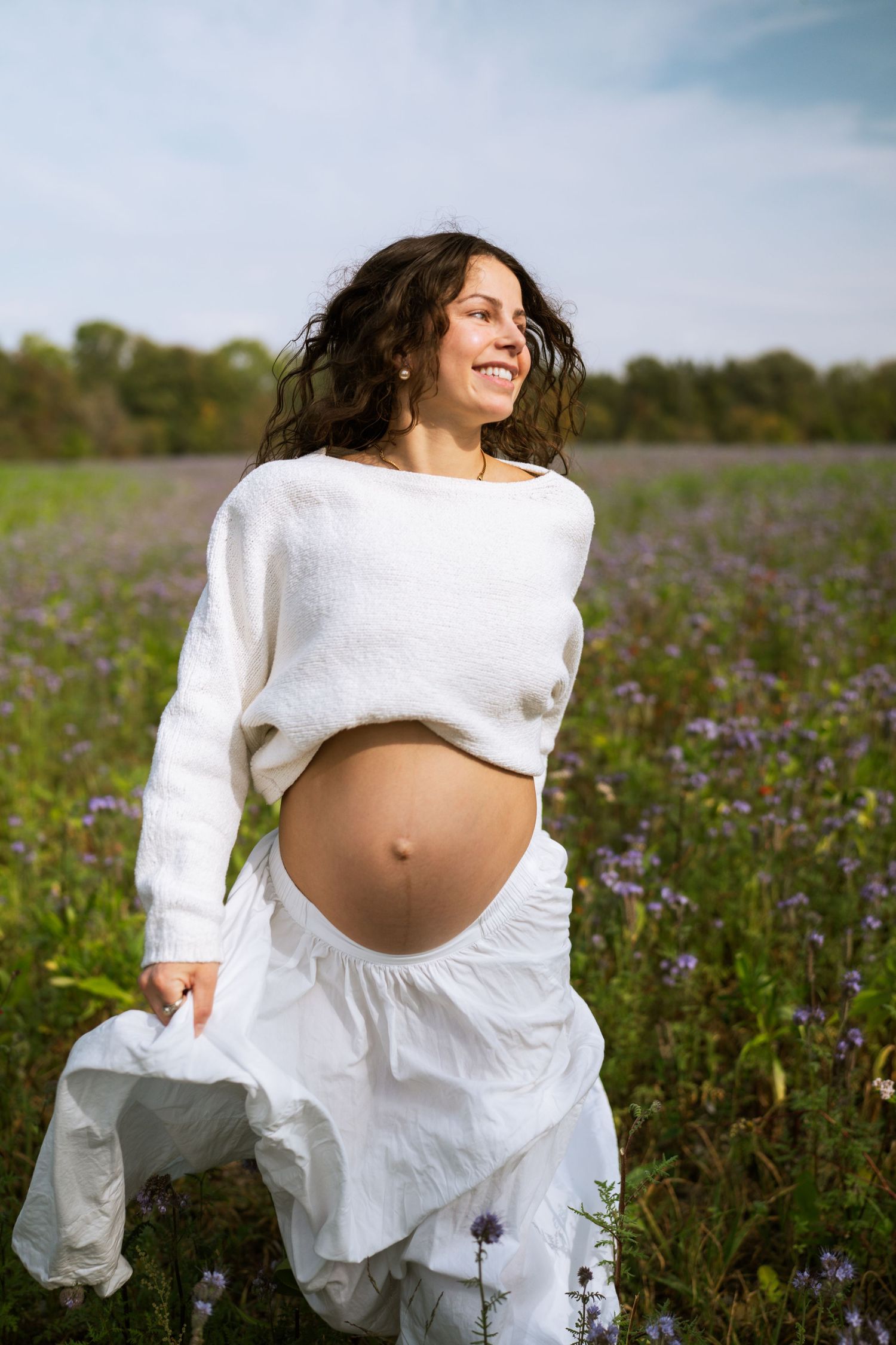 Laughing pregnant woman in a white sweater and flowing skirt runs through a wildflower meadow under a blue sky.