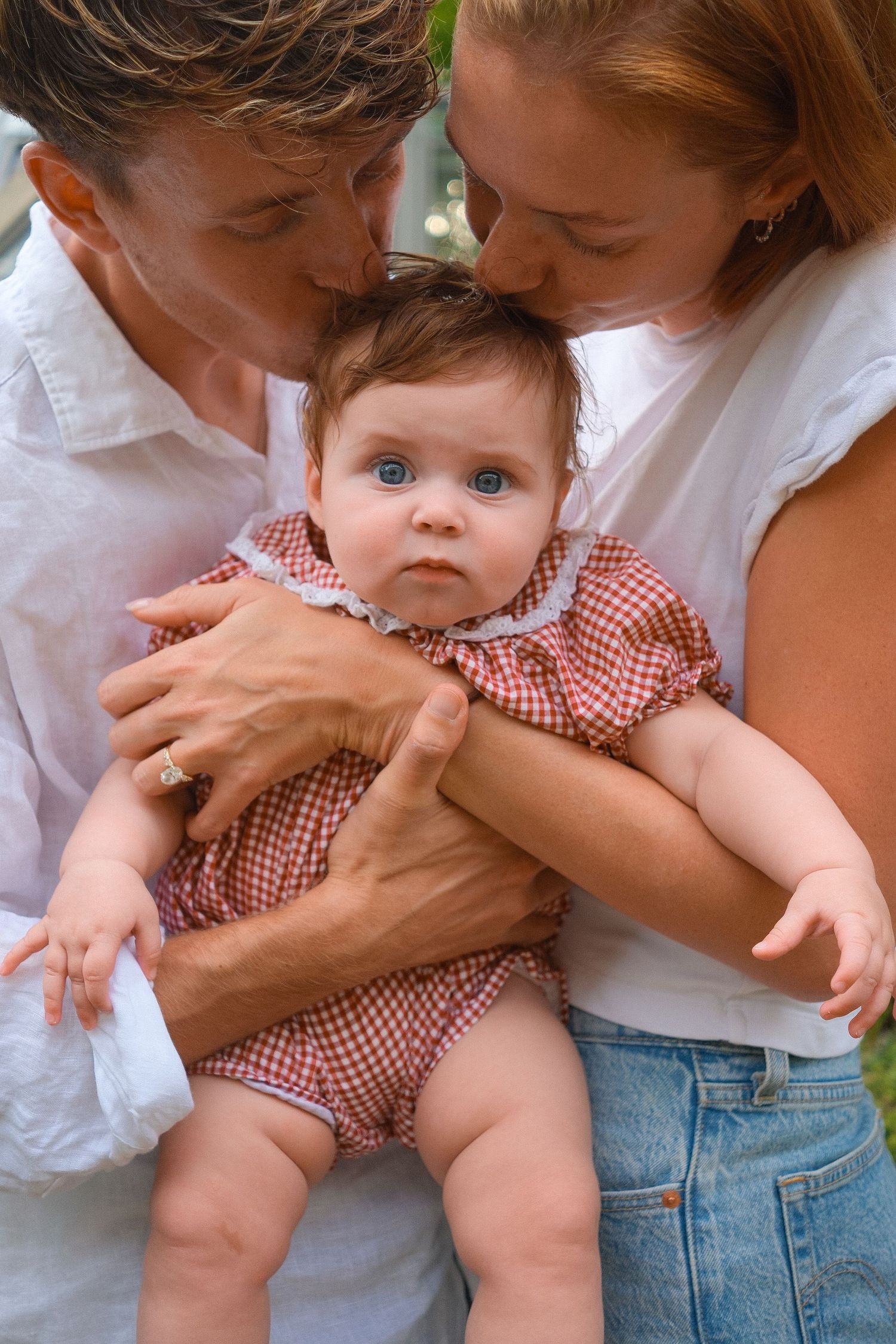 Two parents in white tops kiss the head of a blue-eyed baby girl wearing a pink gingham romper, in a tender family portrait.